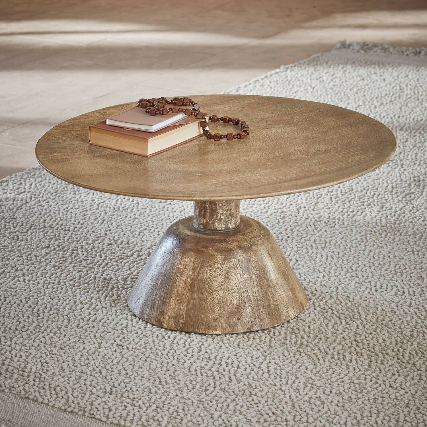 Mesa de centro Ulric Mesa de centro redonda de madera color natural sobre alfombra clara con libros y collar de cuentas