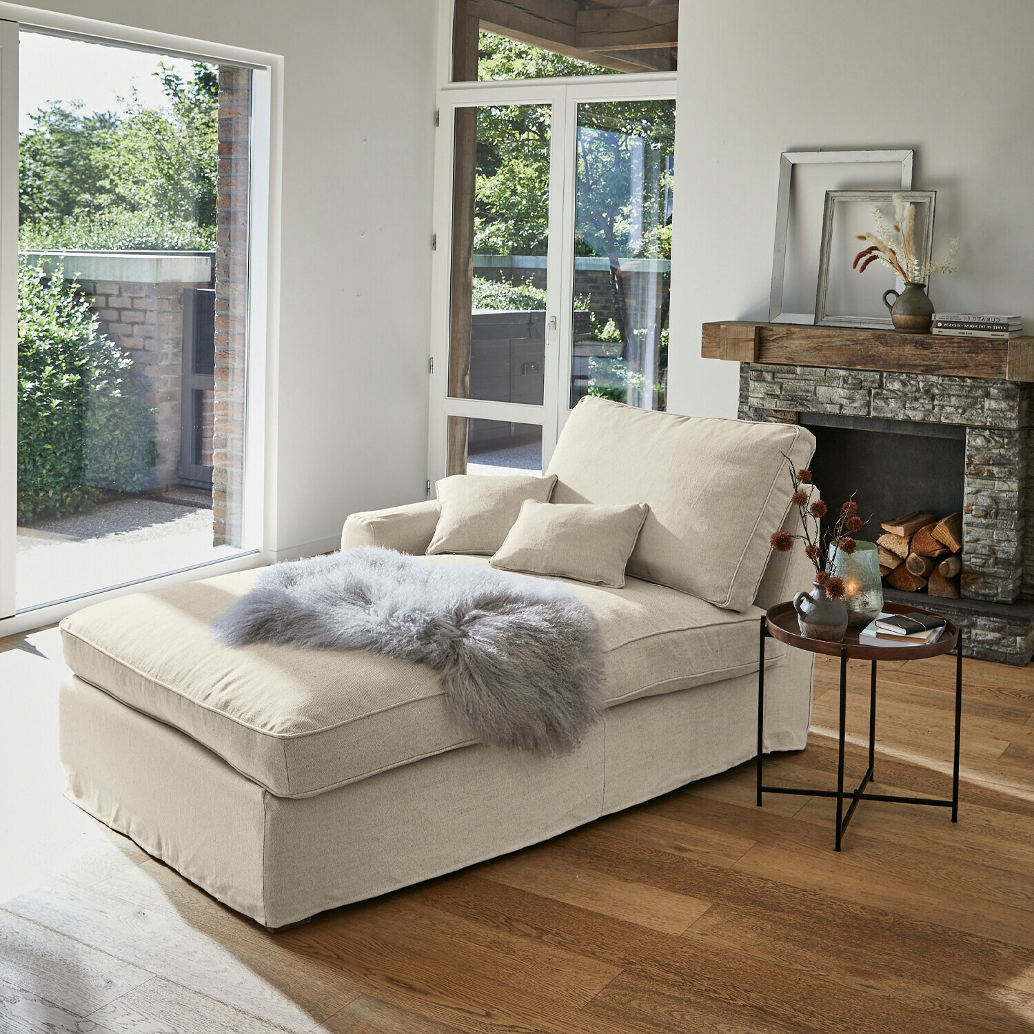 Chaise longue beige de tela con cojines y manta gris peluda en sala luminosa con chimenea