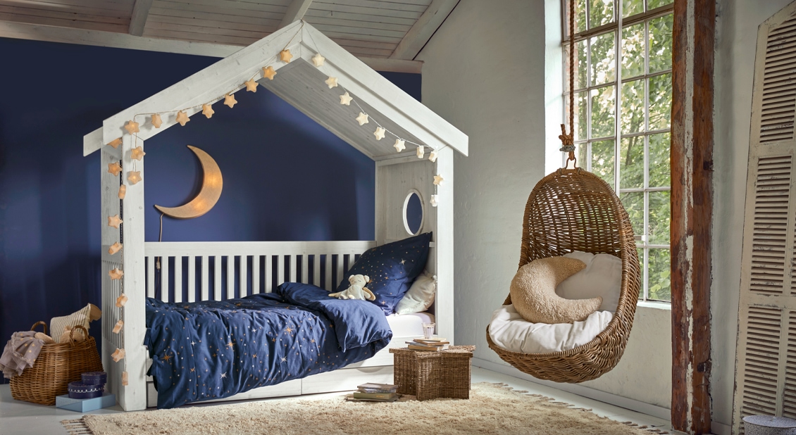 Cama infantil blanca de madera con forma de casita, ropa de cama azul marino y sillón colgante de mimbre en habitación infantil