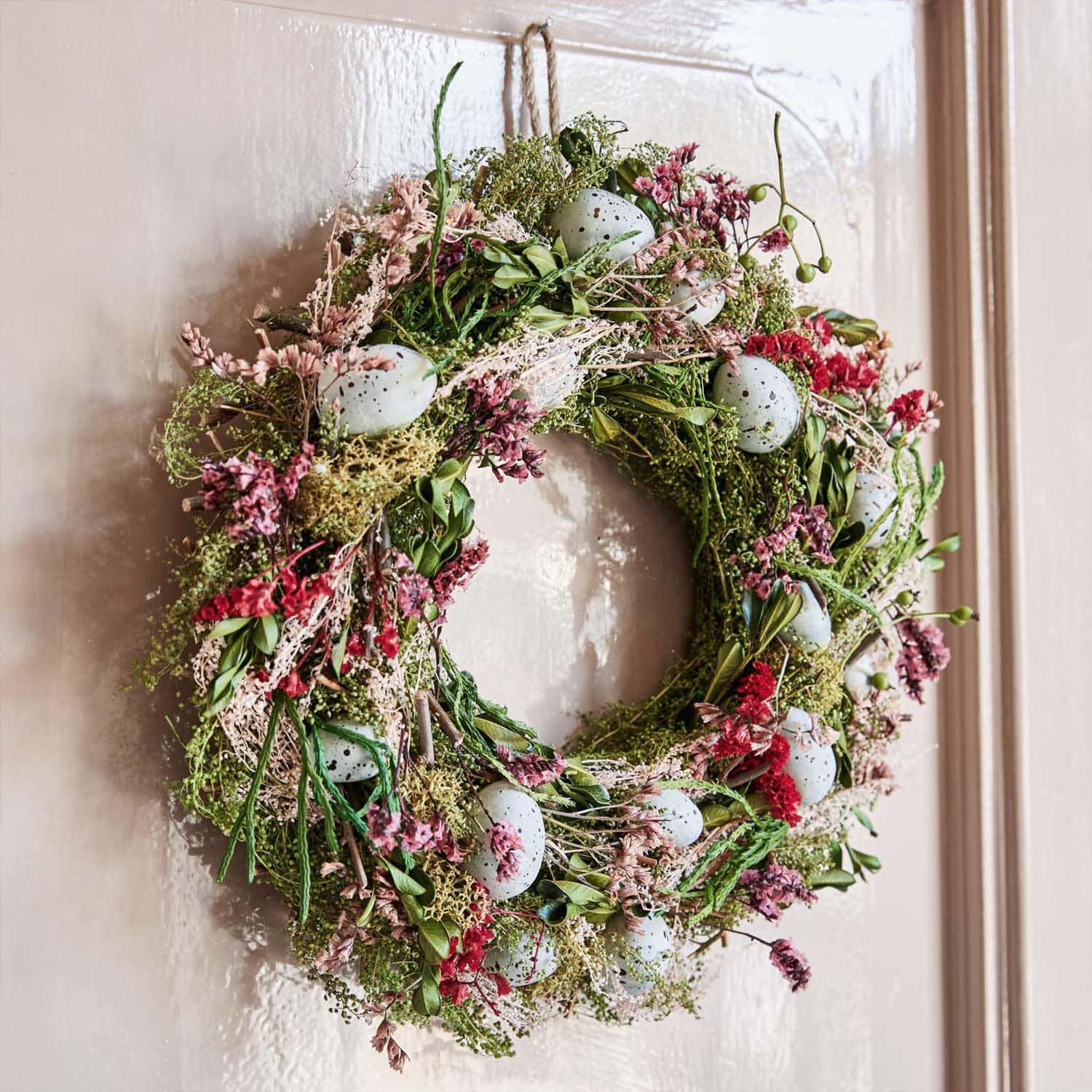 Corona decorativa con ramas verdes, flores rosas y huevos azules artificiales colgada en la puerta