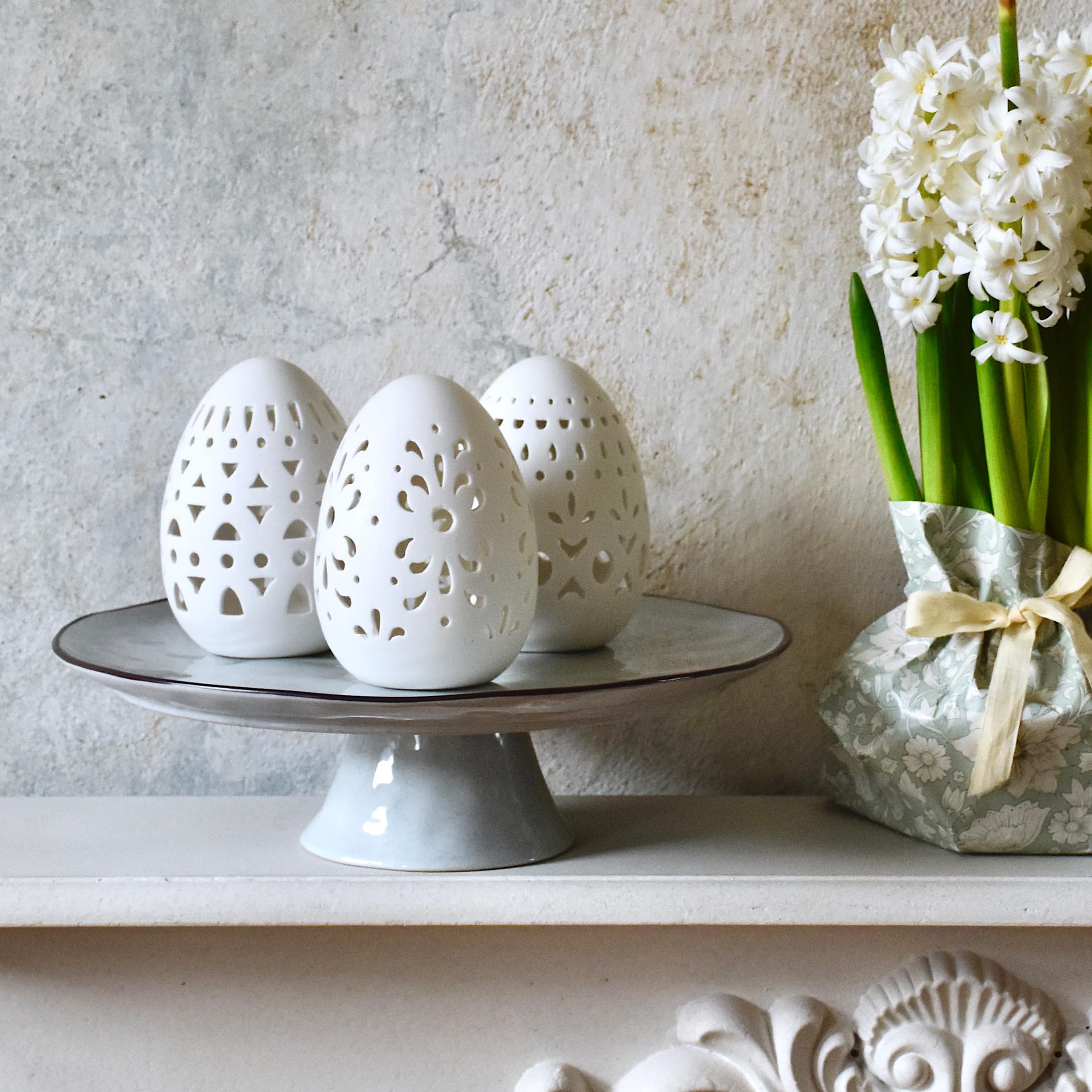Huevos de cerámica blanca con patrones calados sobre plato gris, decoración de Pascua en tonos pastel