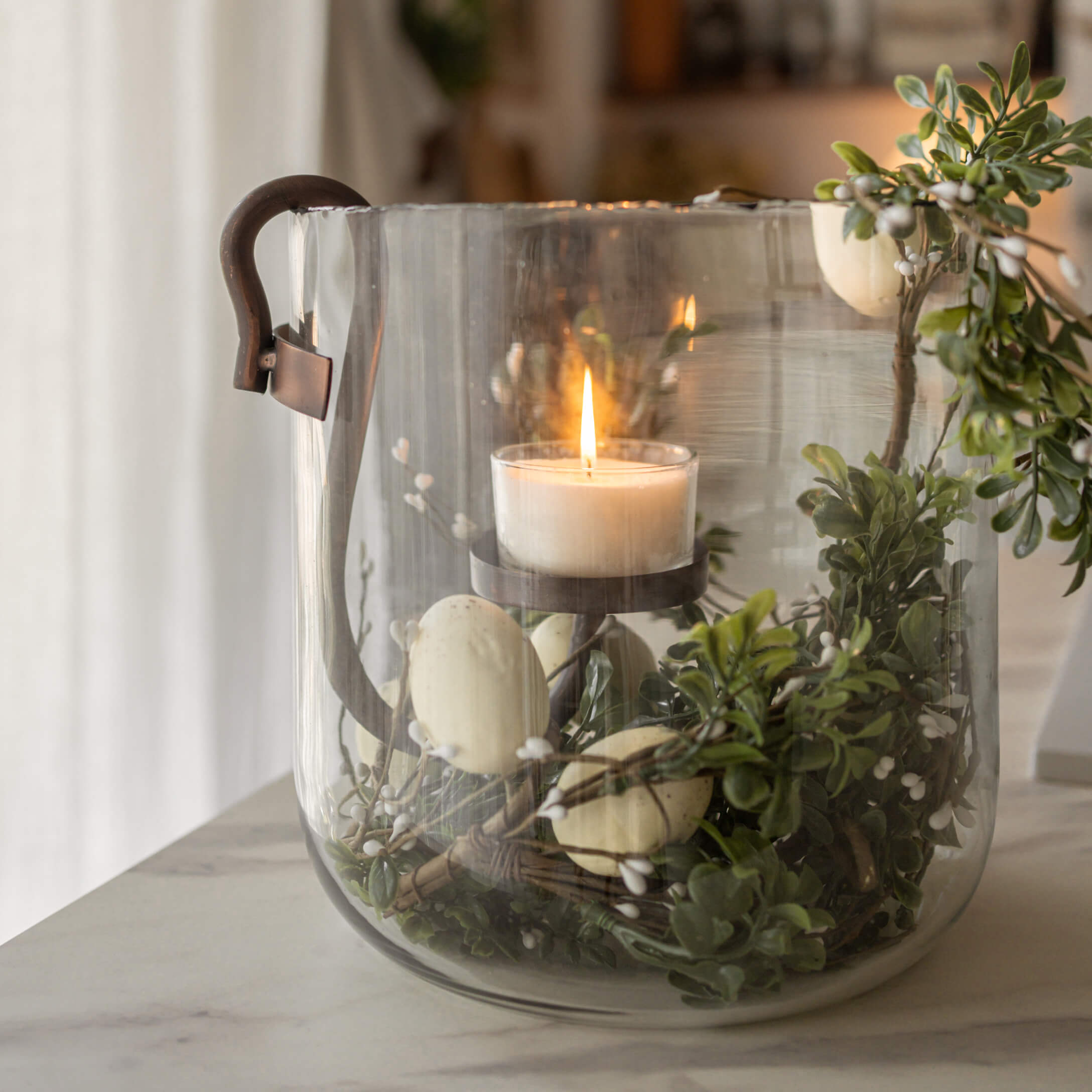 Farol de cristal con asa de metal, vela blanca encendida y decoración verde con frutos blancos