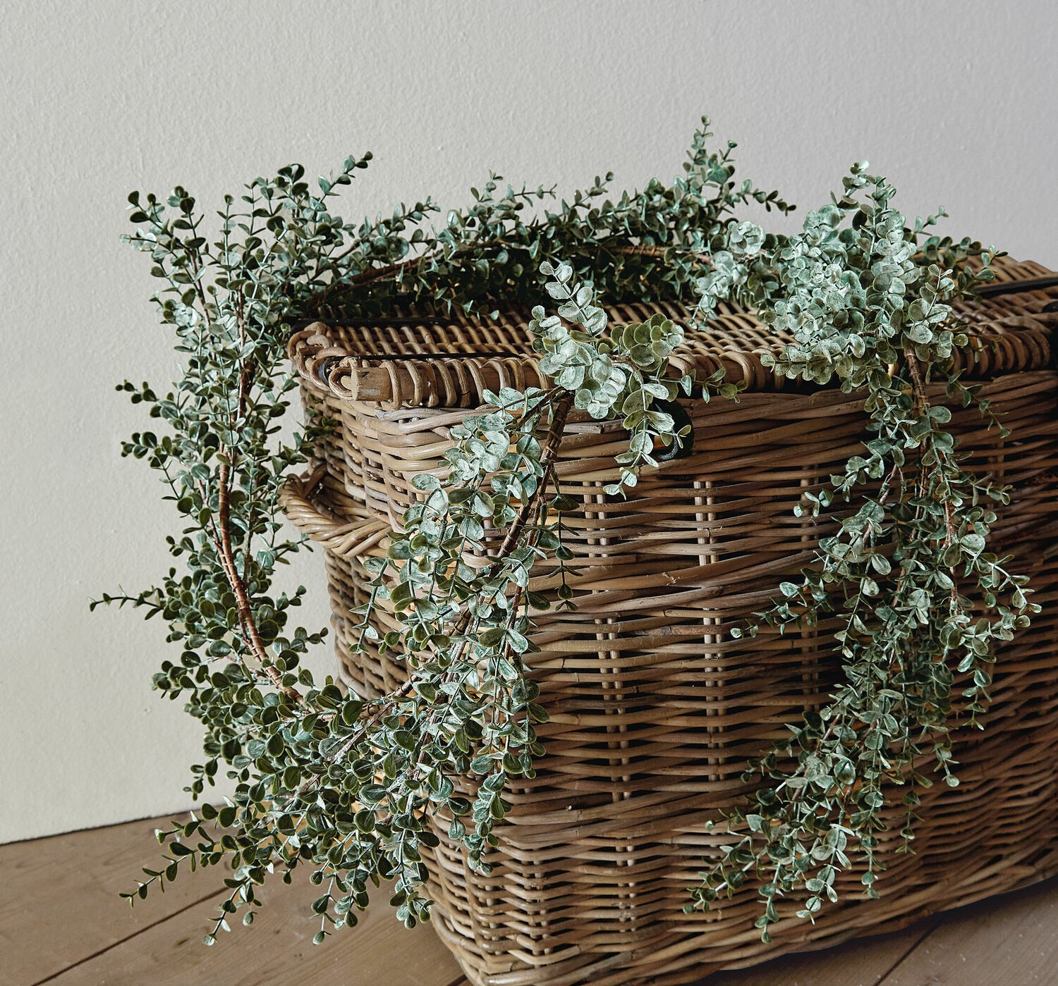 Corona decorativa de hojas verdes de eucalipto sobre cesta de mimbre en suelo de madera