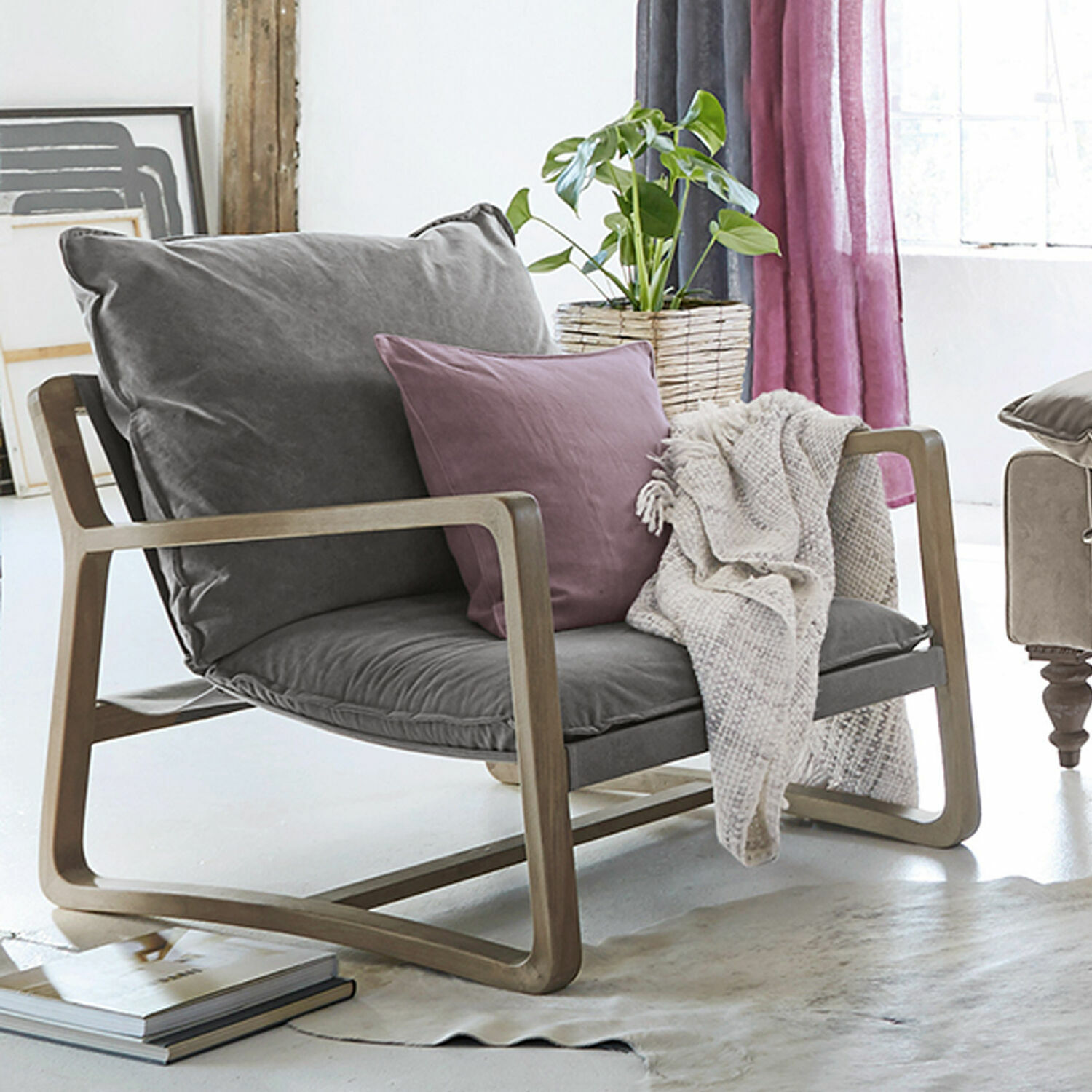 Sillón Jowna Sillón con tapizado gris y estructura de madera, cojín morado y manta beige en sala luminosa