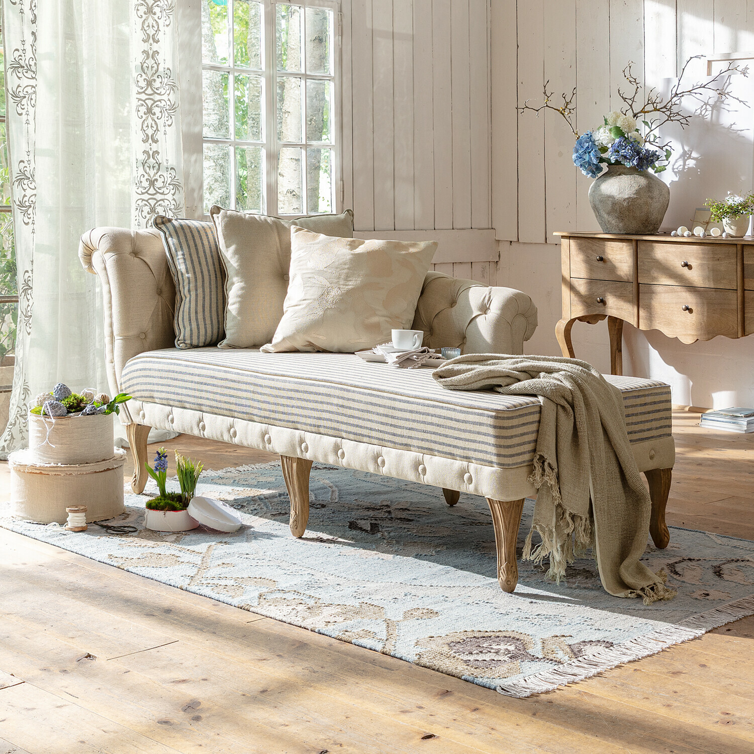 Chaise longue Viviani Sofá beige capitoné con patas de madera y asiento a rayas en salón luminoso con cojines decorativos