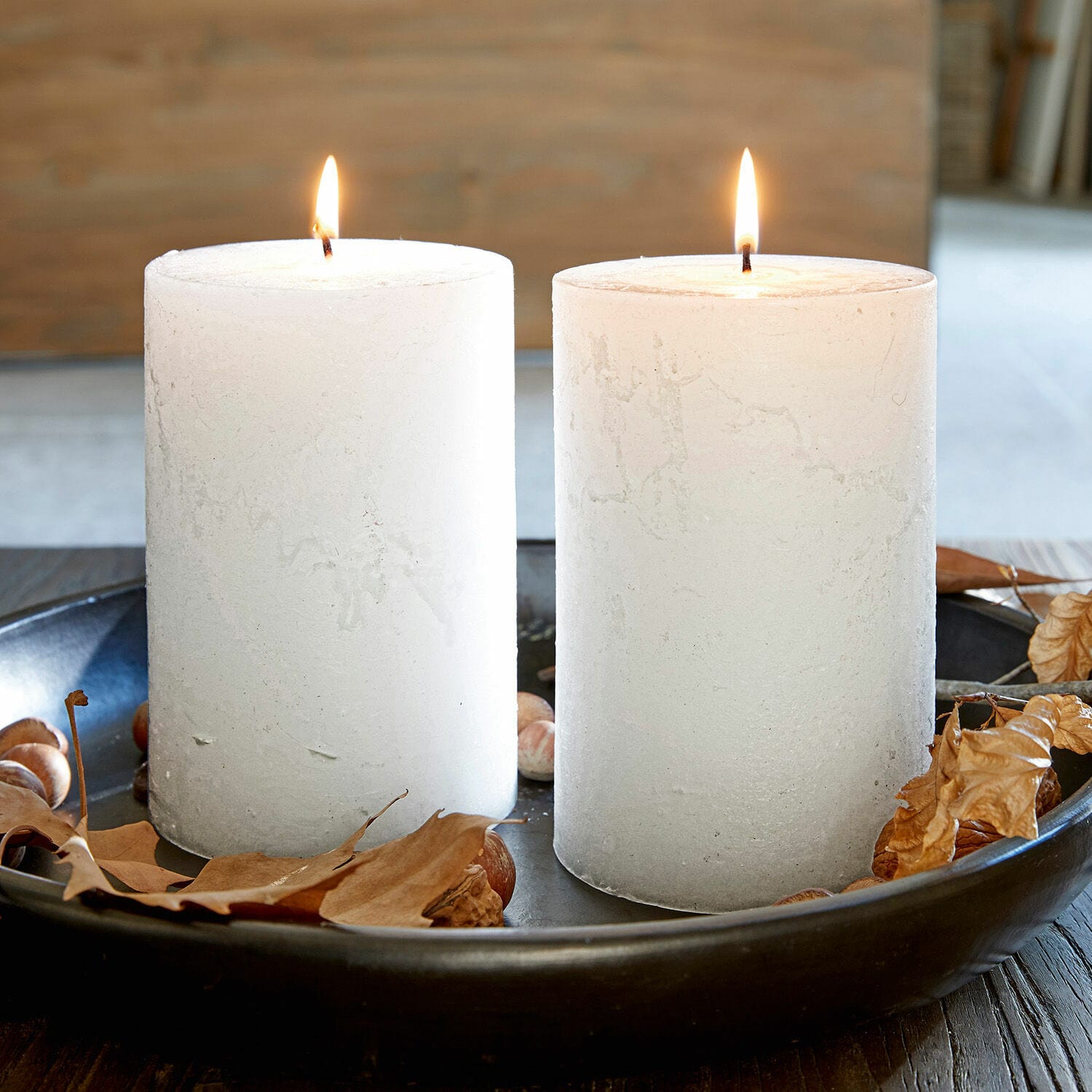 Dos velas blancas de cera en bandeja negra con hojas secas y nueces, decoración otoñal