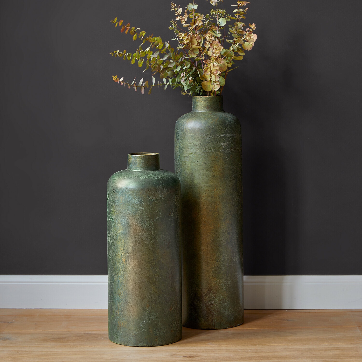 Set de 2 jarrones Darwani Dos jarrones altos de metal verde con hojas secas decorativas sobre suelo de madera