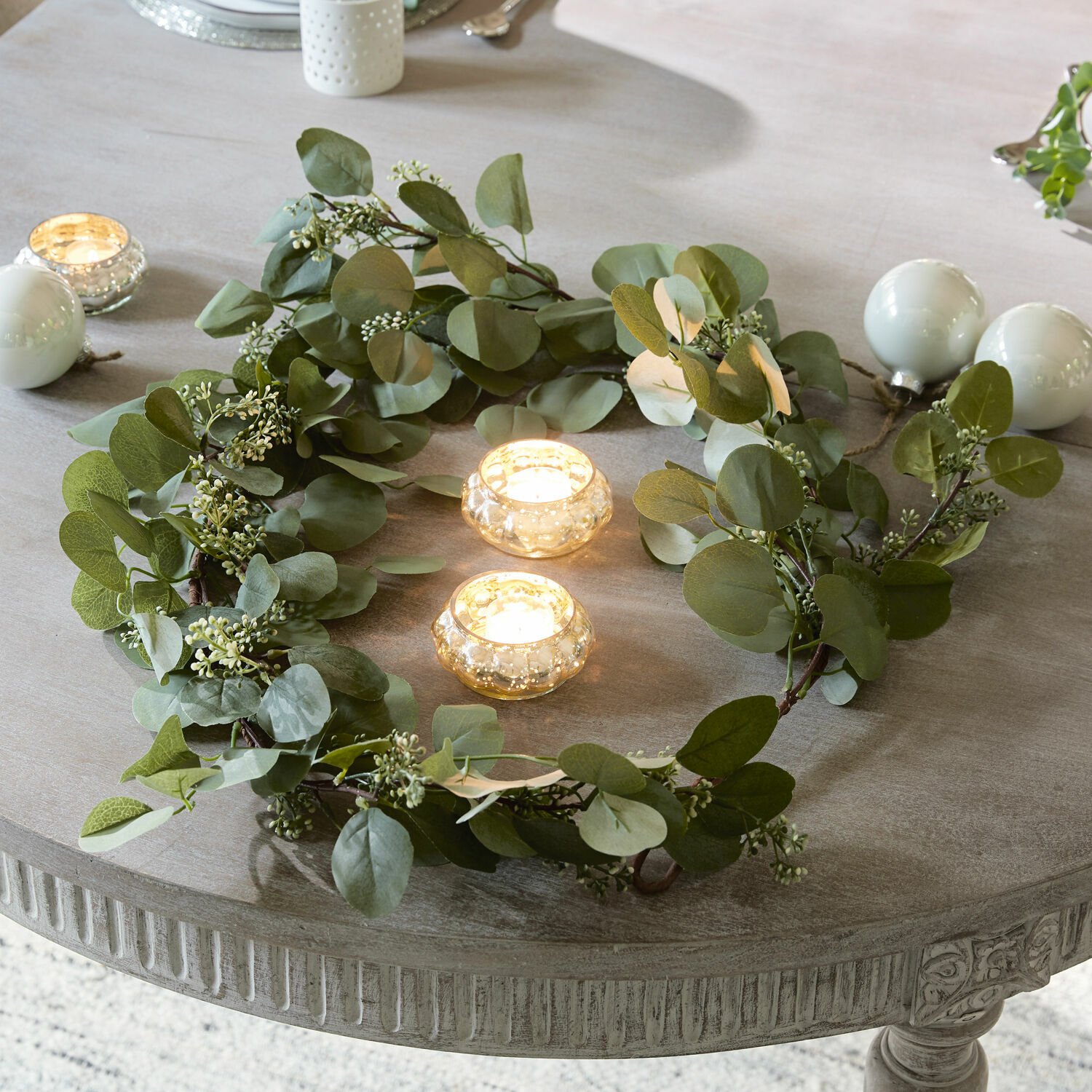 Guirnalda Xantos Corona decorativa de hojas verdes sobre mesa gris con dos velas en portavelas de vidrio