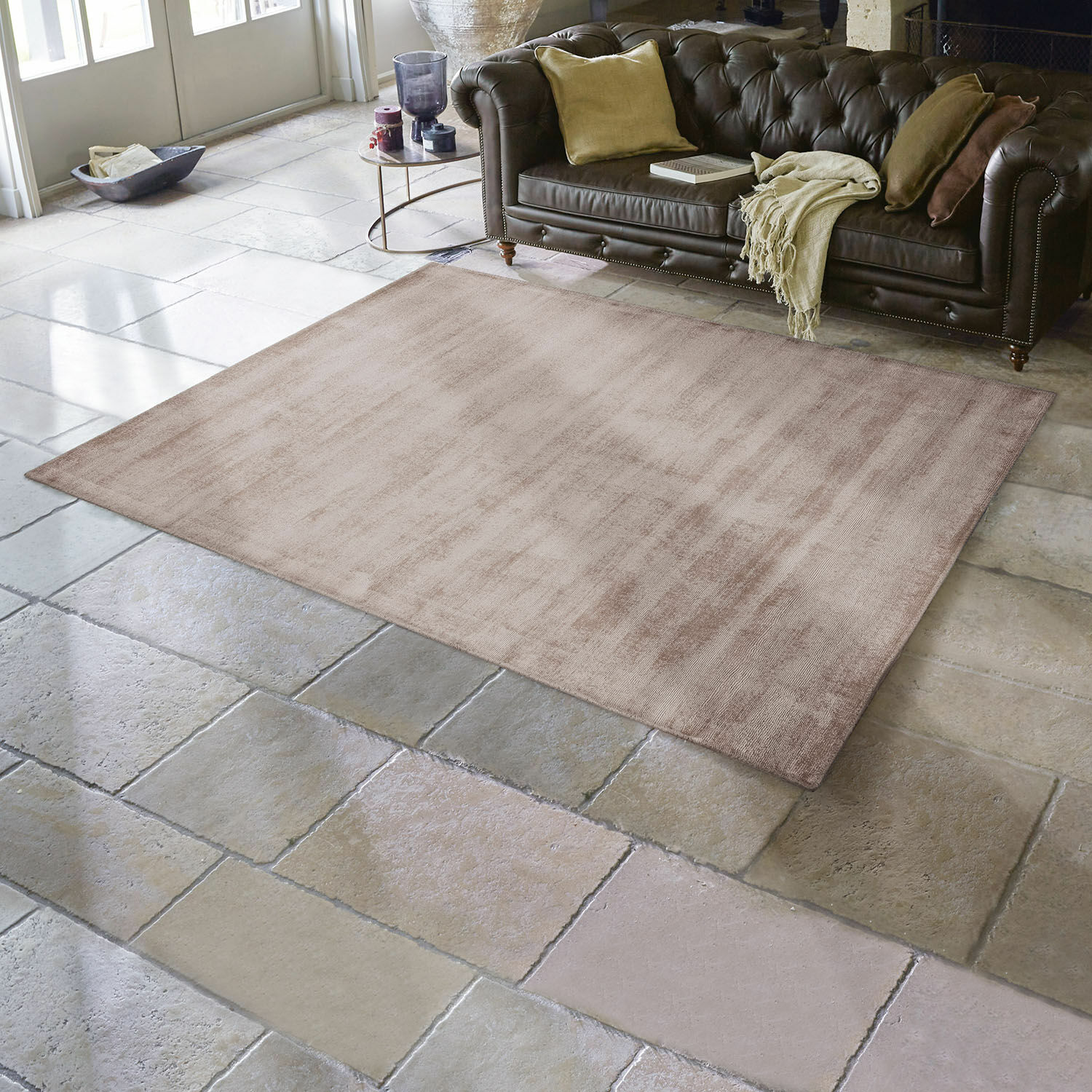 Alfombra Mariselle Alfombra beige de material suave sobre suelo de piedra en salón con sofá de cuero marrón