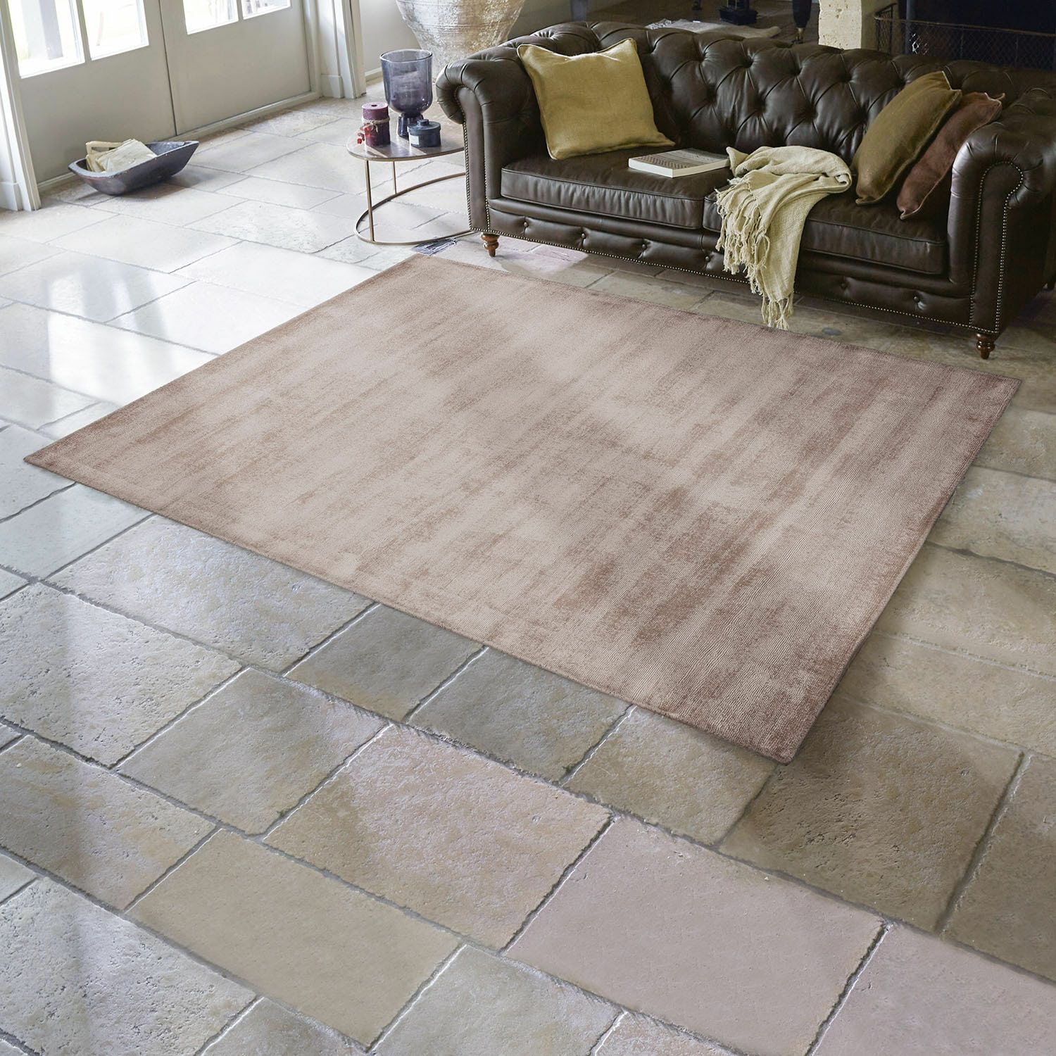Alfombra Mariselle Alfombra beige de material suave sobre suelo de piedra en salón con sofá de cuero marrón