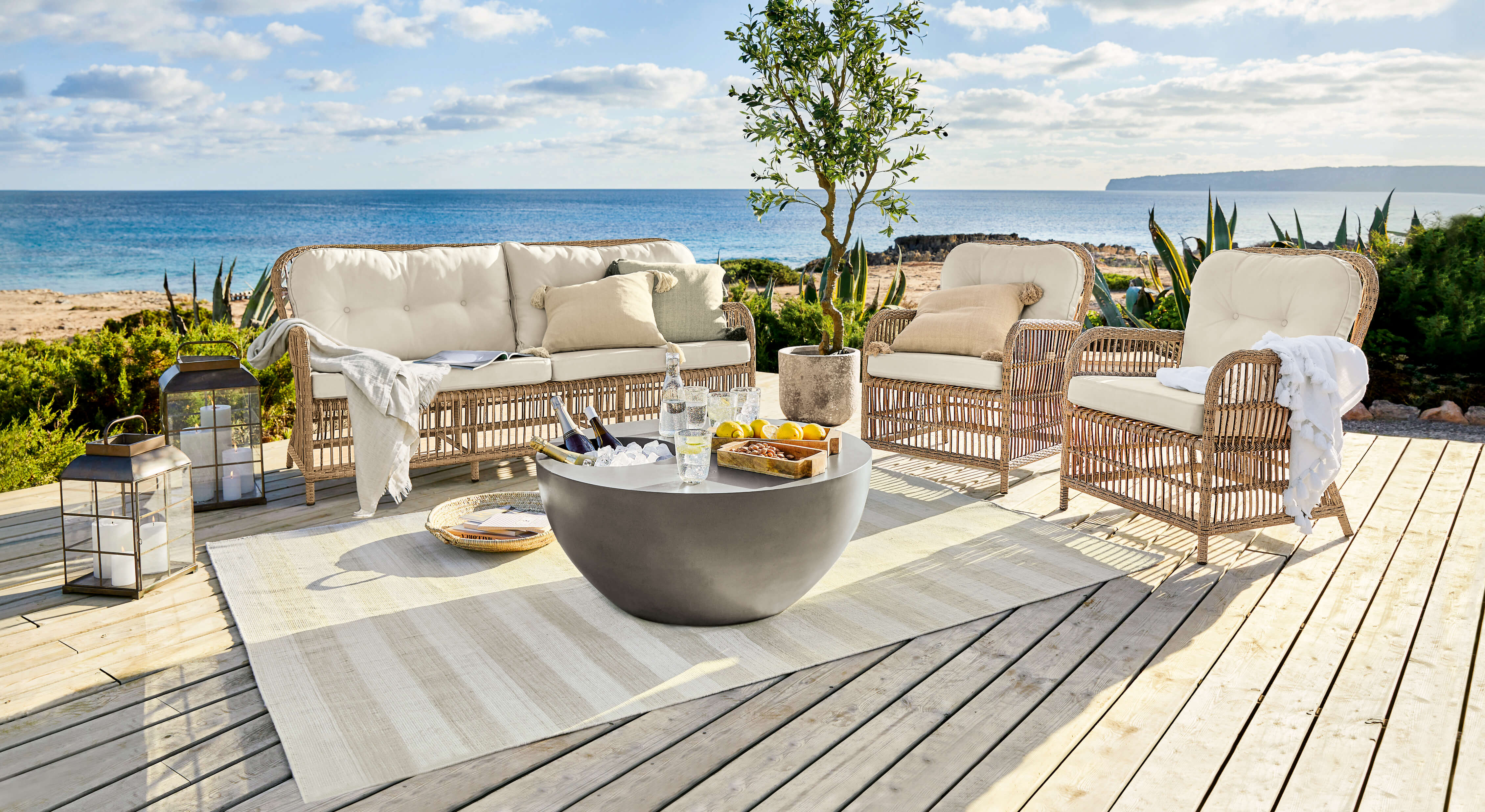Conjunto de muebles de jardín de ratán con cojines crema en terraza de madera con vista al mar