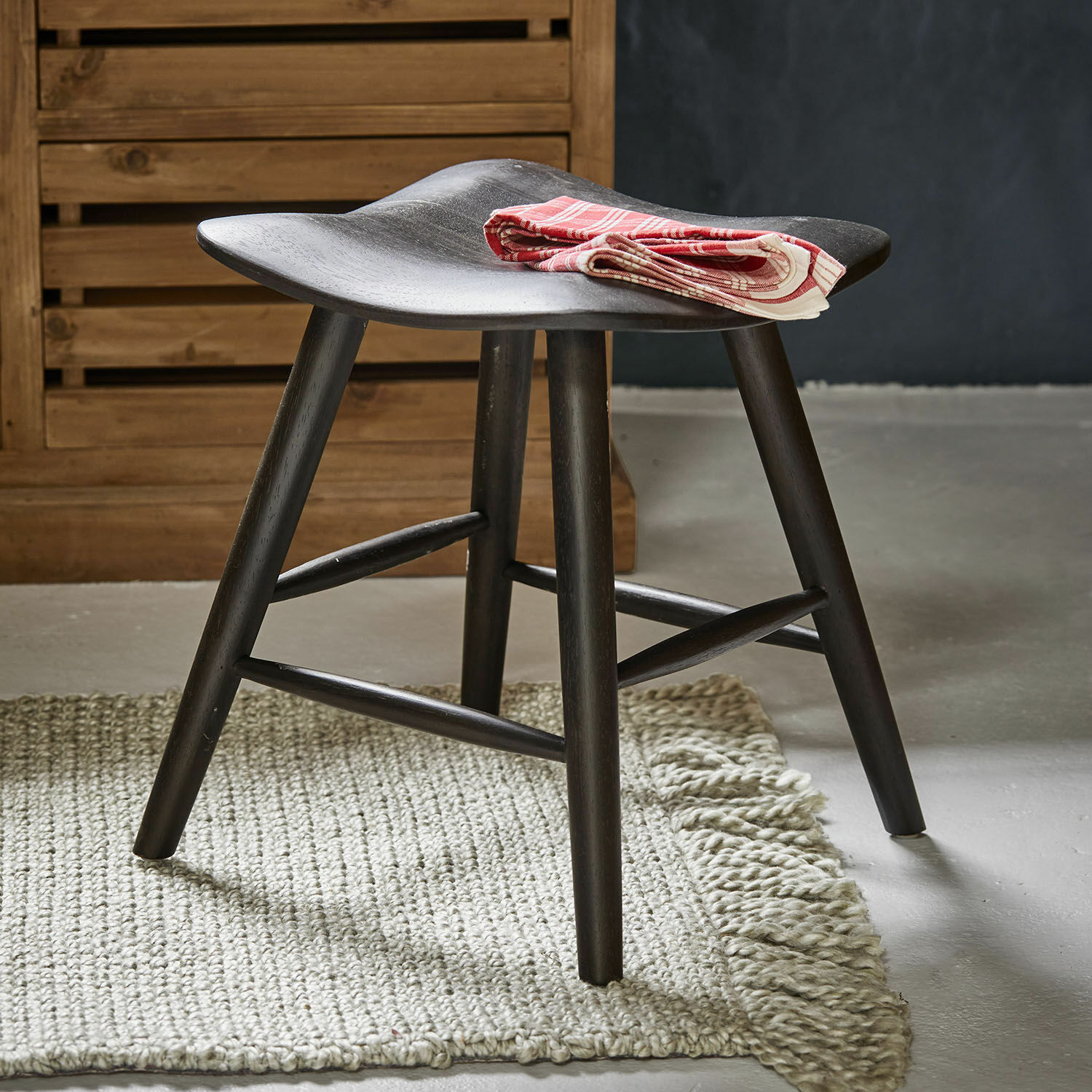 Taburete Voiláren Taburete de madera negra con asiento curvado sobre alfombra clara, paño decorativo rojo doblado