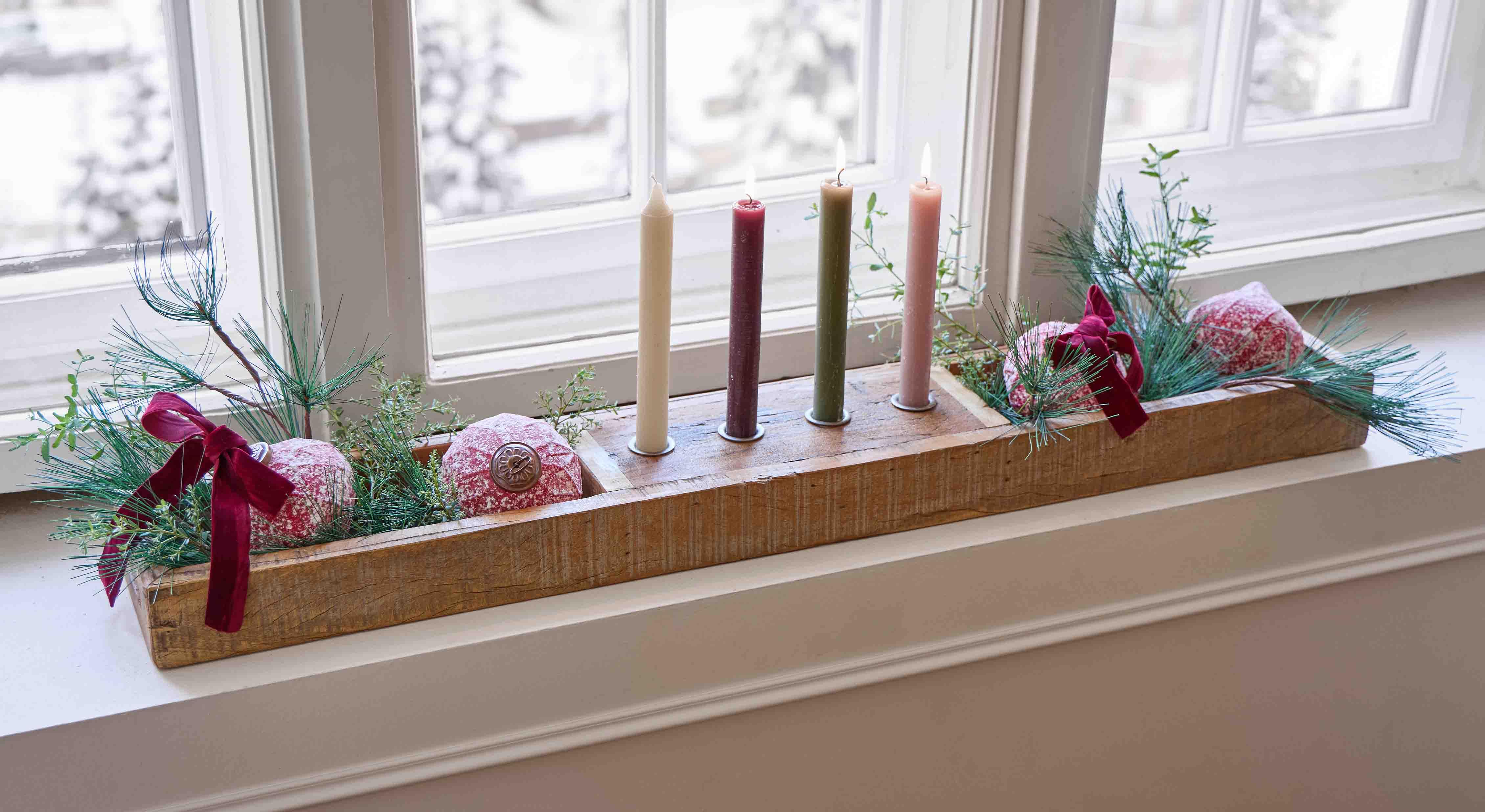 Base de madera con cuatro velas de colores y decoraciones verdes y rojas en el alféizar de la ventana
