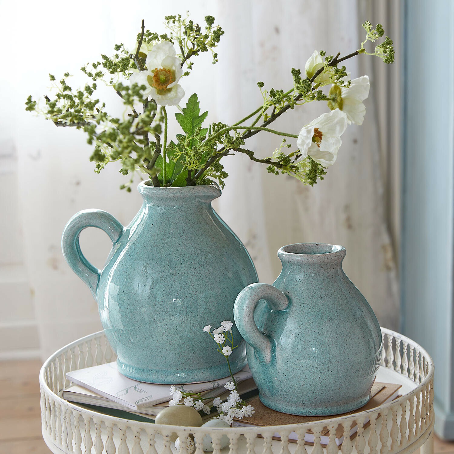 Dos jarras de cerámica turquesa con asas sobre mesa metálica blanca con flores y libros