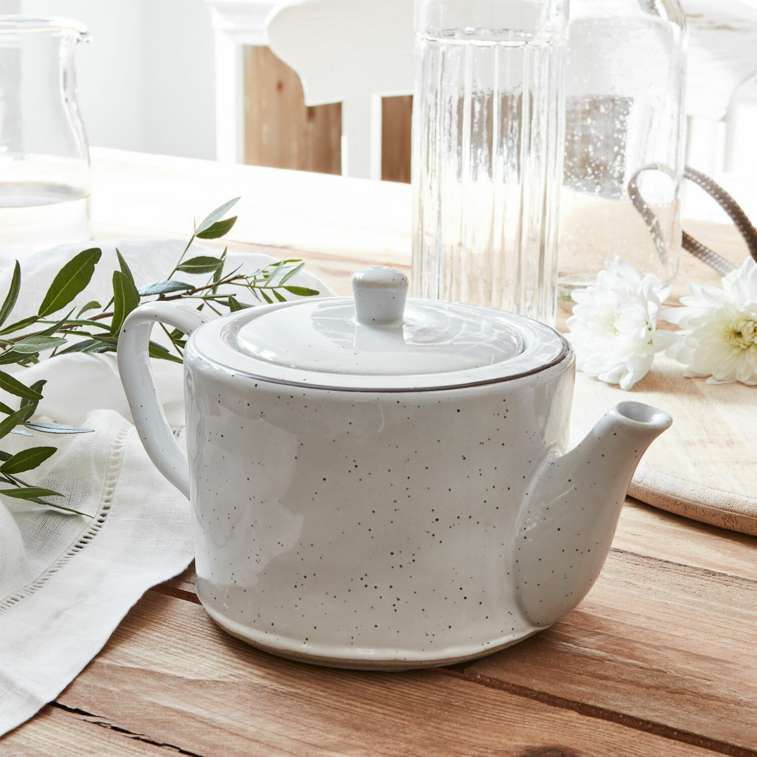 Tetera de cerámica blanca con tapa sobre mesa de madera con flores blancas y hojas verdes