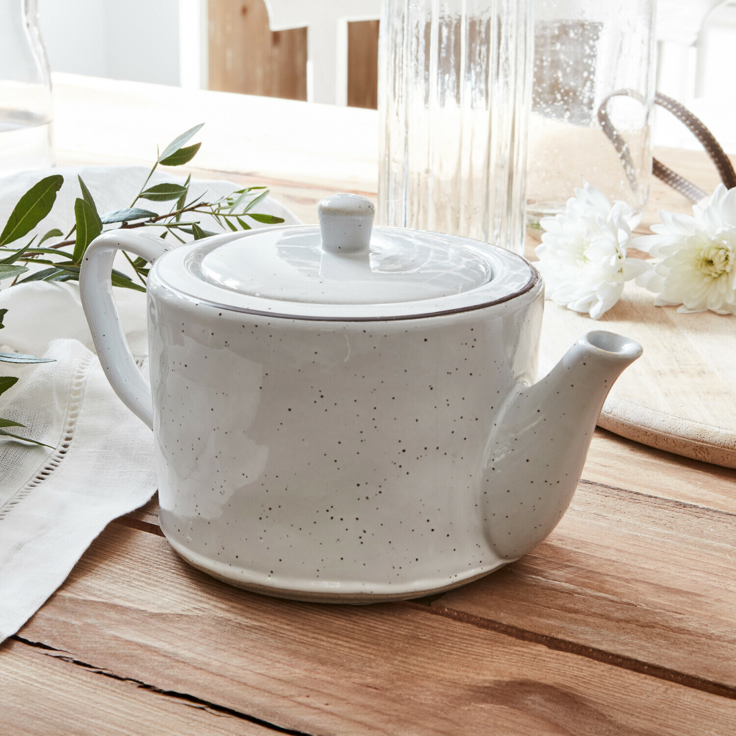 Tetera Biarré Tetera de cerámica blanca con tapa sobre mesa de madera con flores blancas y hojas verdes