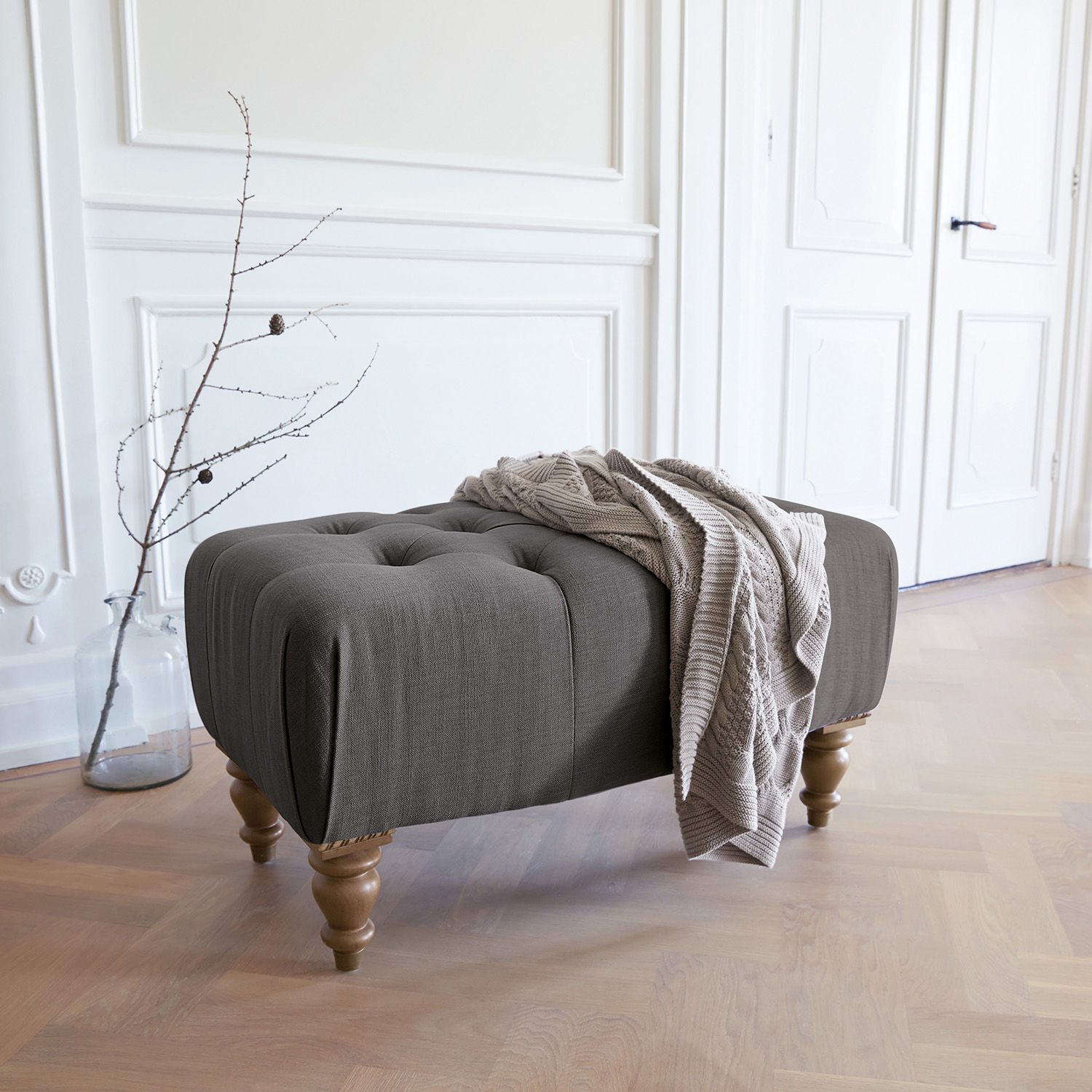 Taburete Durance Puf tapizado gris con patas de madera y manta beige en interior luminoso