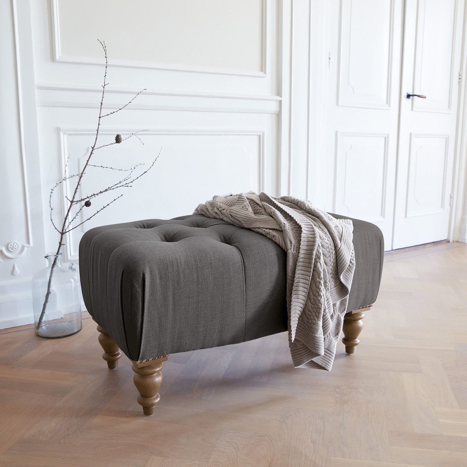 Taburete Durance Puf tapizado gris con patas de madera y manta beige en interior luminoso