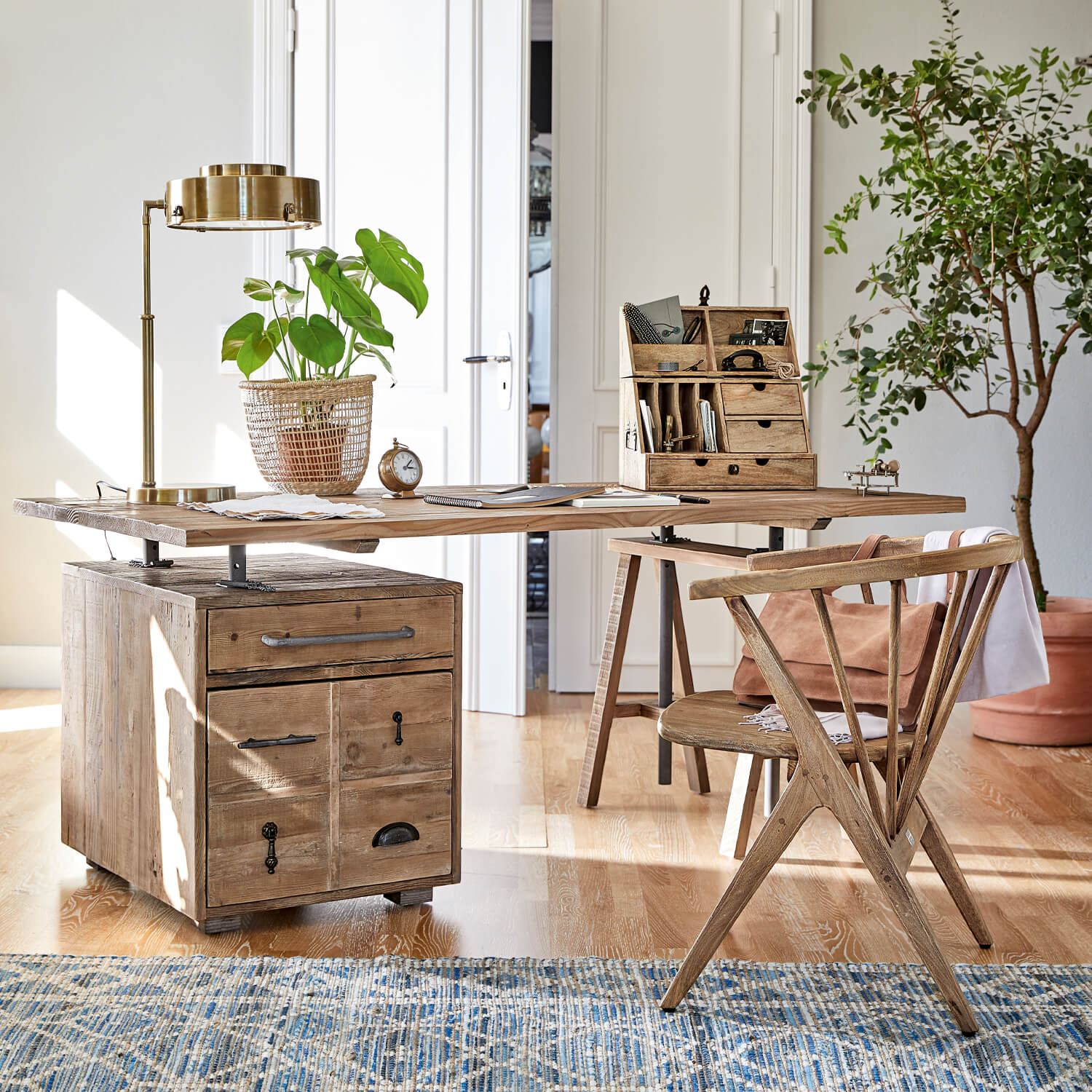 Escritorio de madera con cajones y tiradores metálicos, lámpara dorada y planta en cesta de mimbre