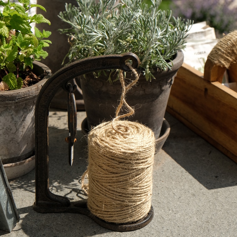 Bobina de cuerda de yute natural en soporte metálico junto a macetas con hierbas para jardinería