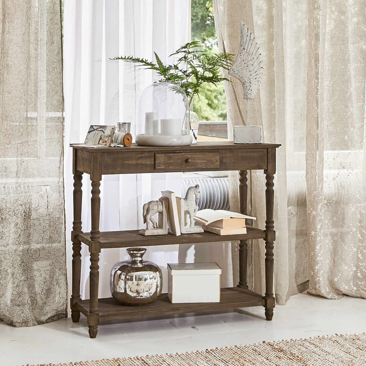 Consola Roubaix Mesa de madera marrón con cajón y estantes, decorada con libros y jarrones en interior luminoso