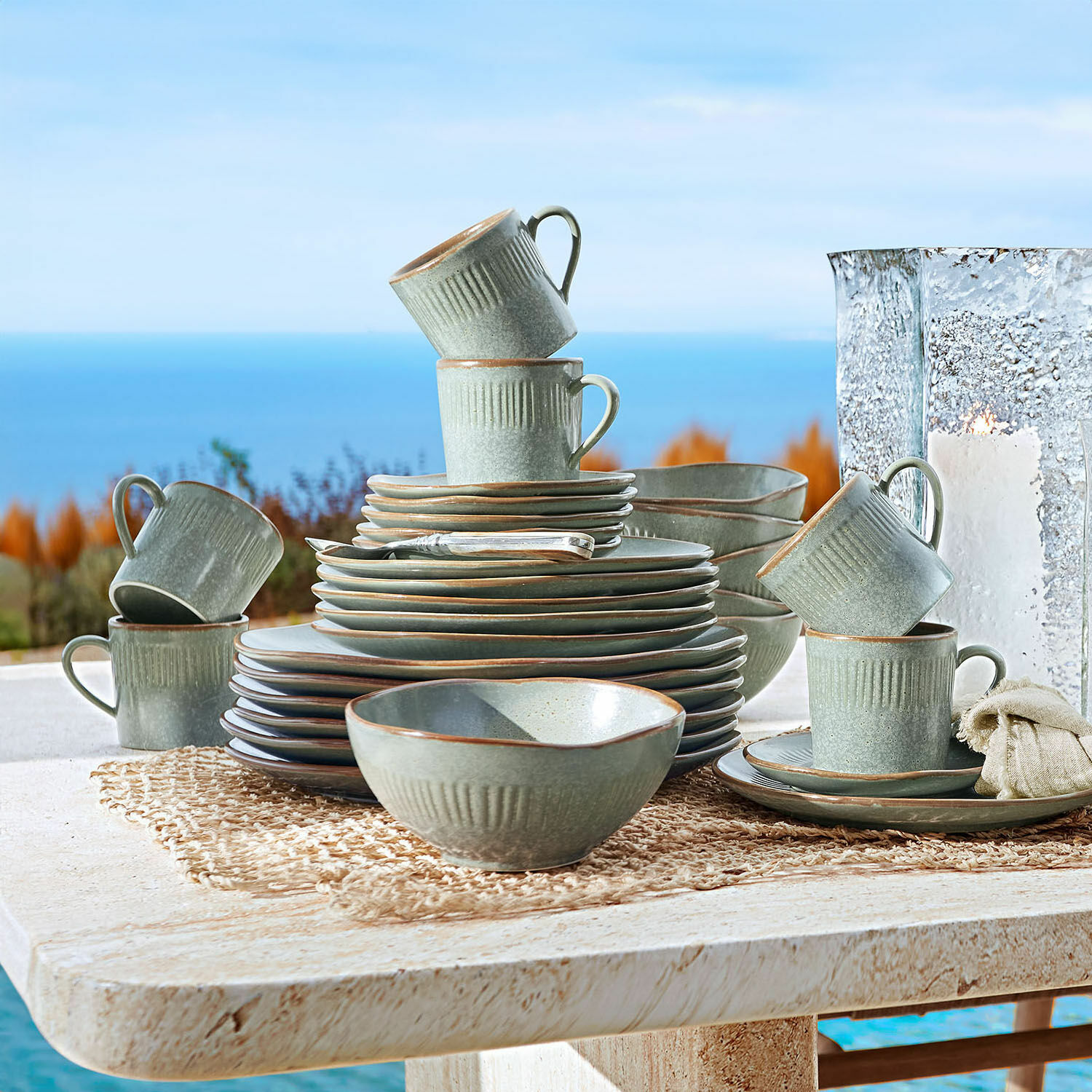 Set de 30 piezas de vajilla Périers Juego de vajilla de cerámica gris con tazas y platos apilados en mesa exterior con vista al mar