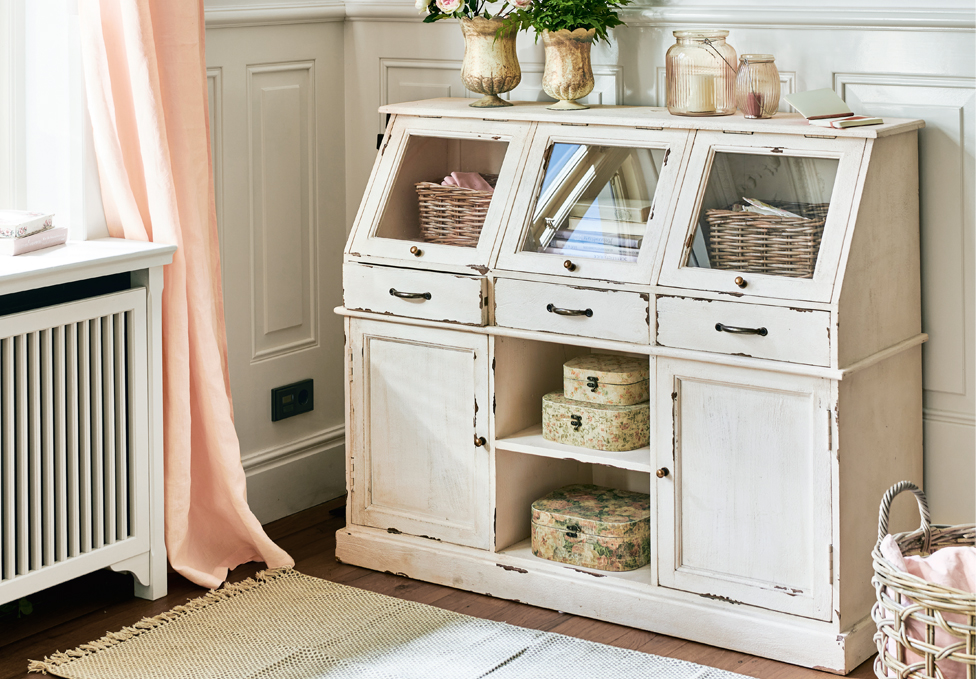 Aparador de madera blanca estilo shabby chic con cajones de cristal y decoración en habitación luminosa.