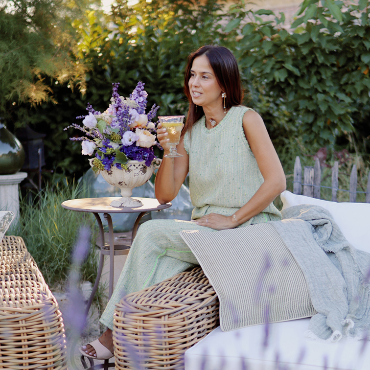 Mujer con vestido verde sentada en mueble de mimbre en jardín con ramo de flores y copa de vino