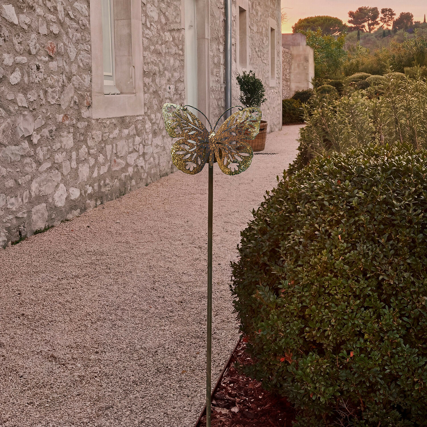 Decoración de jardín metálica en forma de mariposa color latón frente a muro de piedra y arbustos verdes