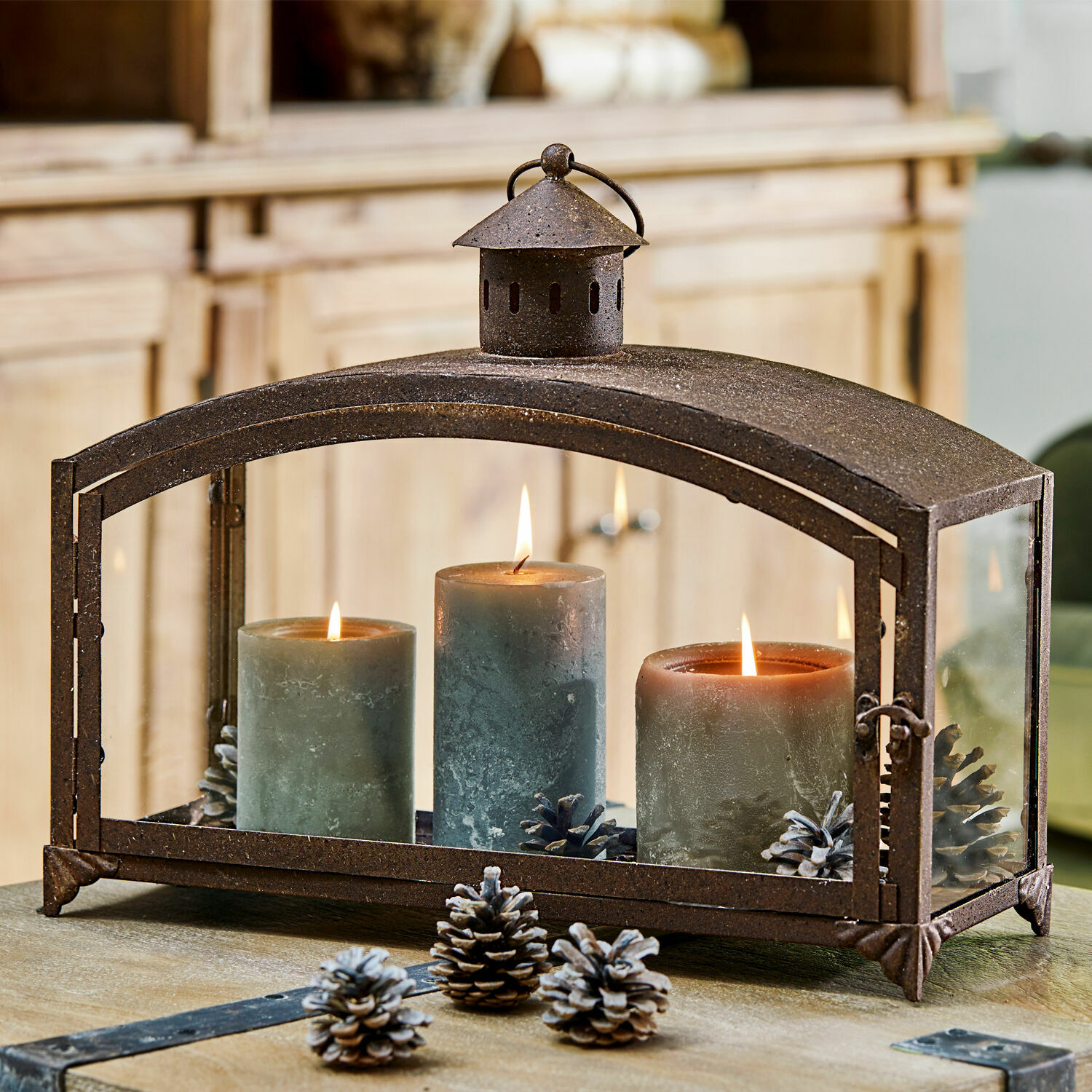 Portavelas Miora Farol de metal con tres velas verdes y piñas sobre mesa de madera, decoración para interiores