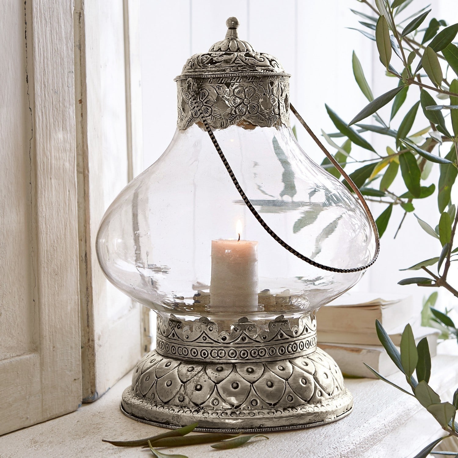 Farol Kaela Linterna de vidrio con base y tapa metálica plateada con diseño floral, vela encendida en el interior