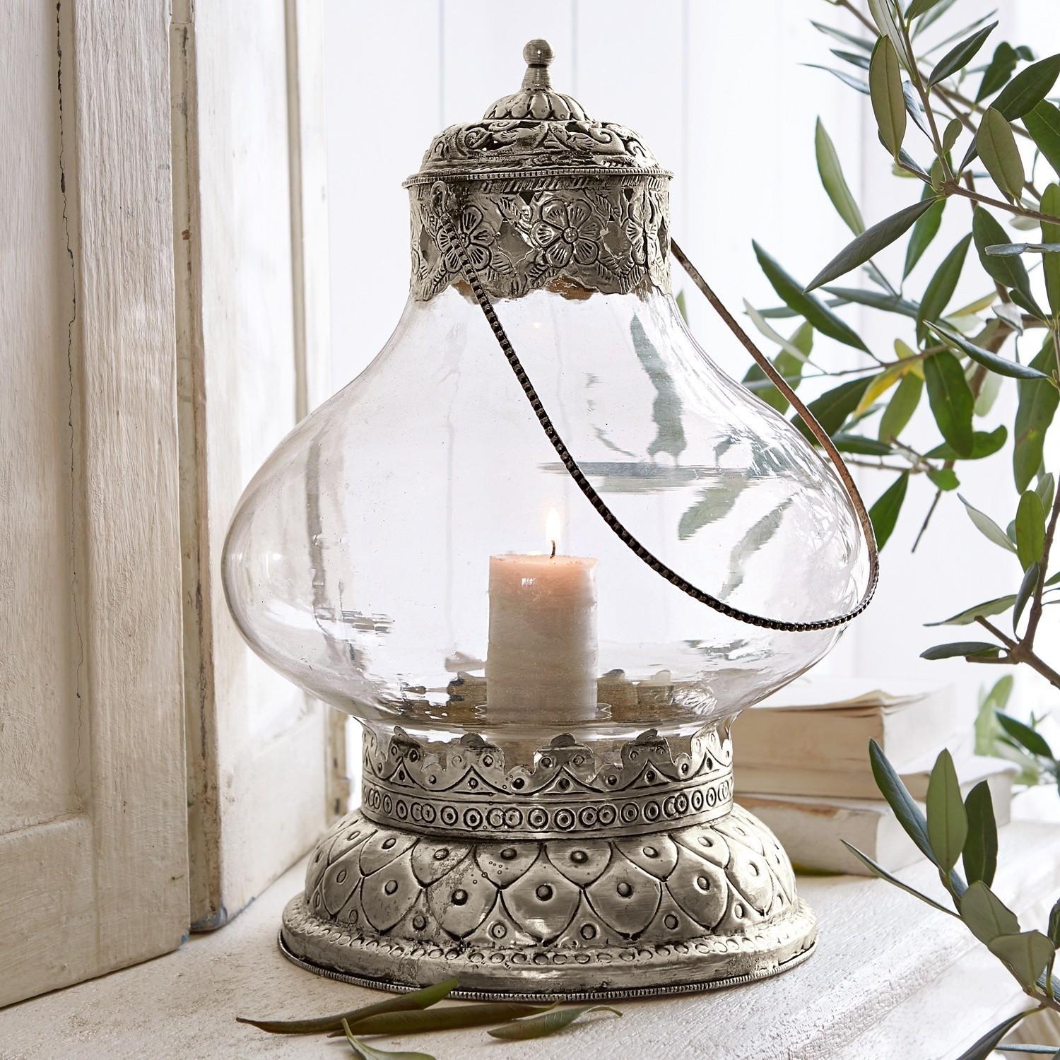 Farol Kaela Linterna de vidrio con base y tapa metálica plateada con diseño floral, vela encendida en el interior