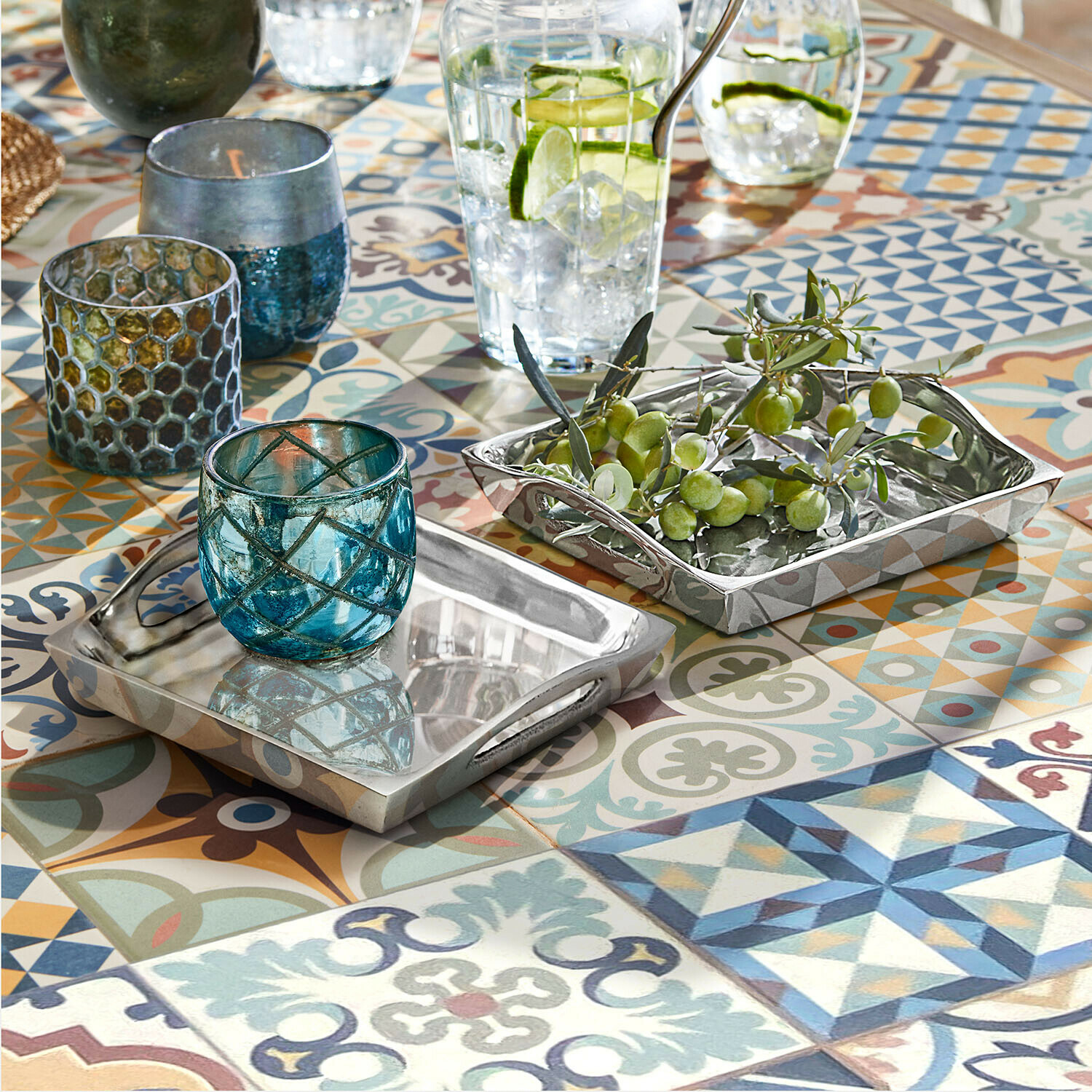 Bandejas metálicas plateadas reversibles sobre mesa con mosaico colorido, portavelas de vidrio azul y aceitunas verdes
