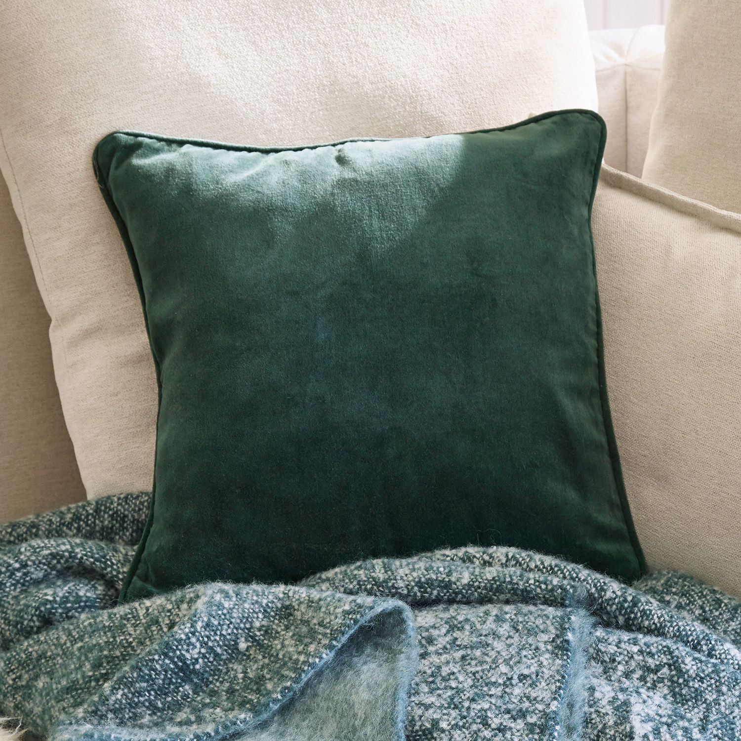 Cojín decorativo verde de terciopelo sobre sofá beige con manta suave al fondo