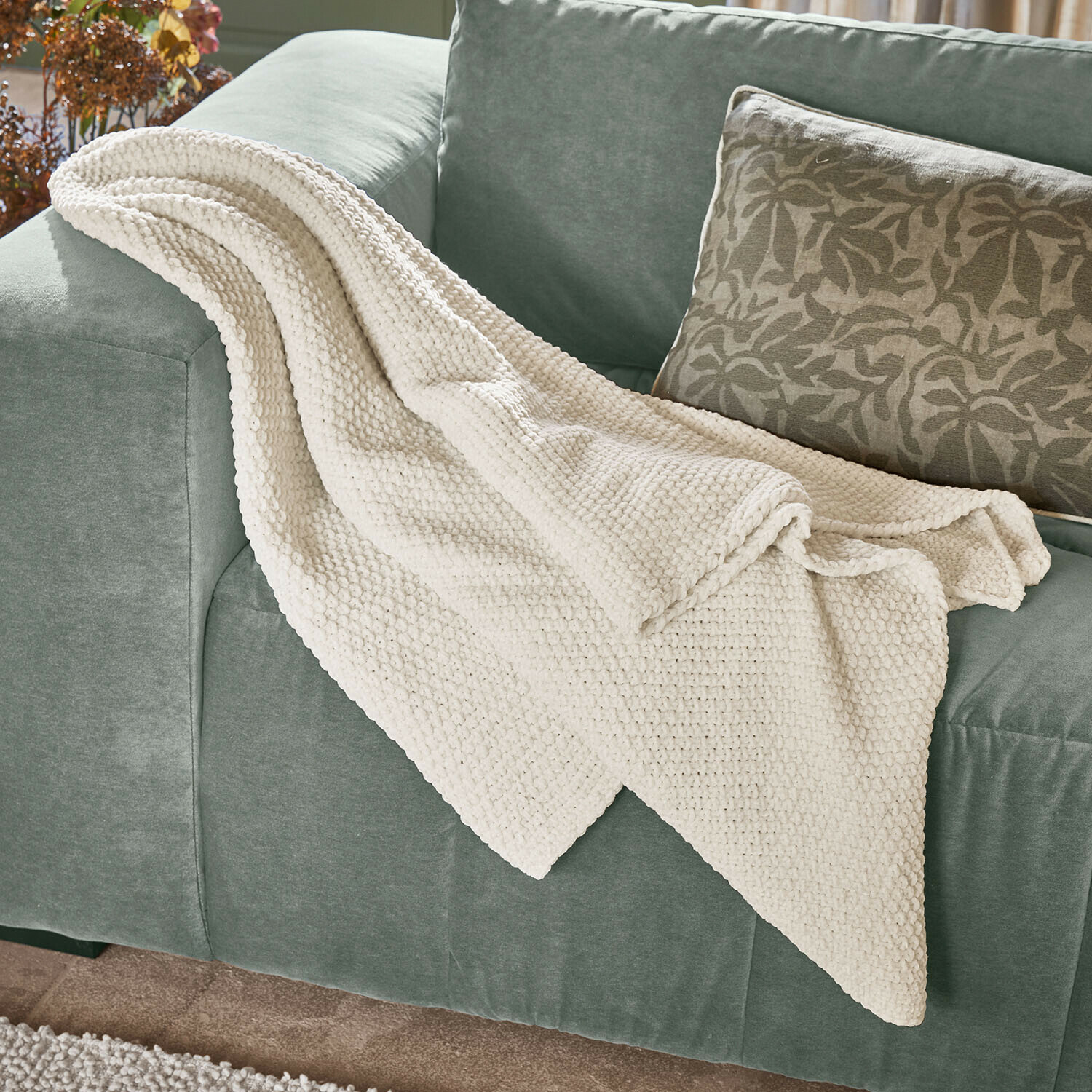 Plaid Linnet Manta cremosa de punto grueso sobre sofá verde, ideal para salón y decoración de interiores