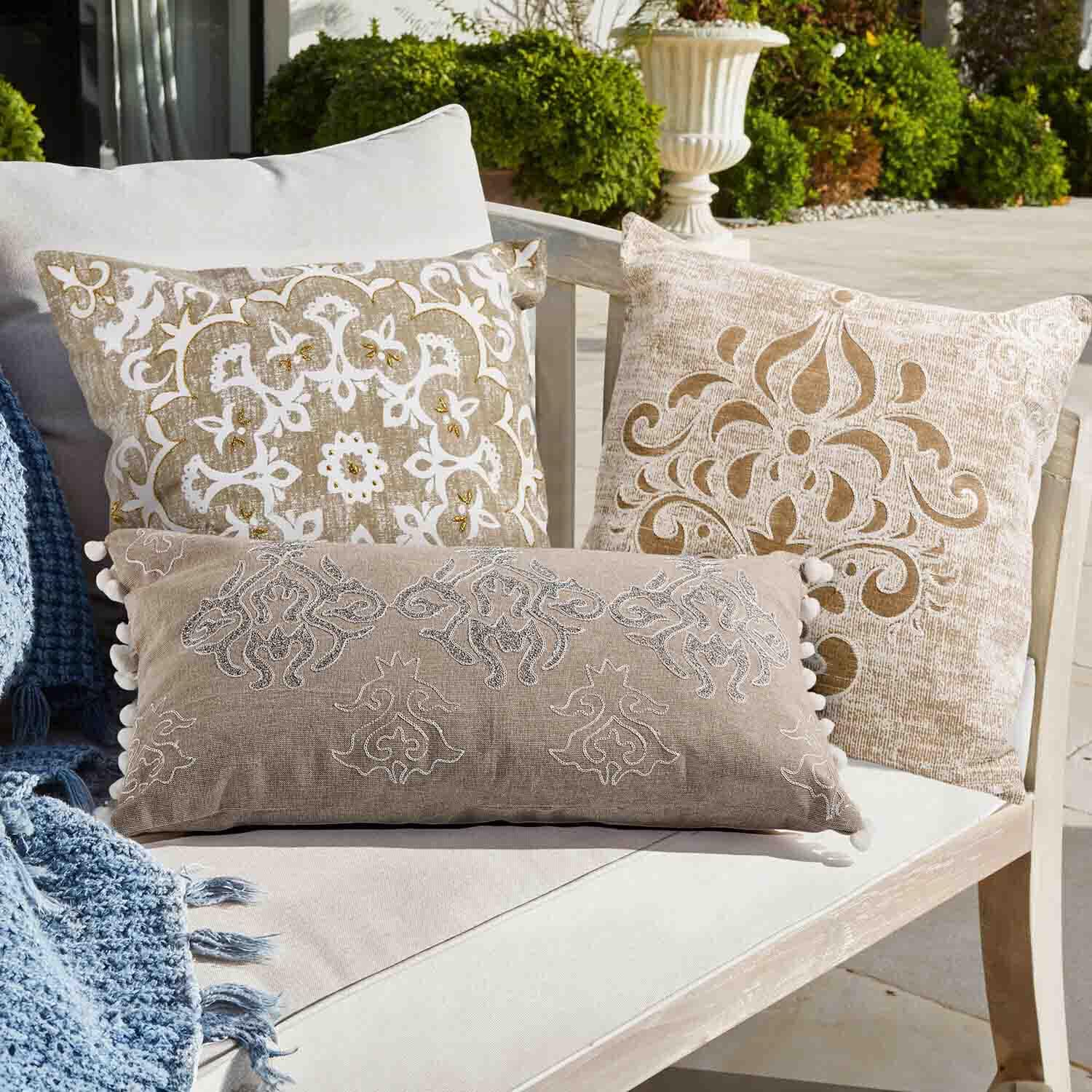 Conjunto de tres cojines decorativos beige y blanco con patrones sobre sofá claro en jardín