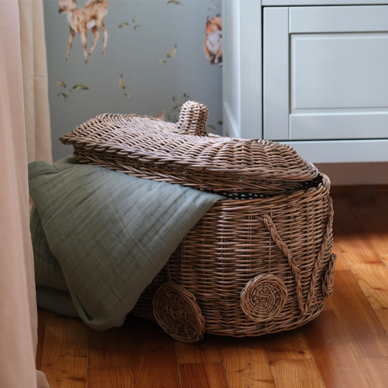 Cesta de mimbre con forma de coche y manta verde sobre suelo de madera en habitación infantil