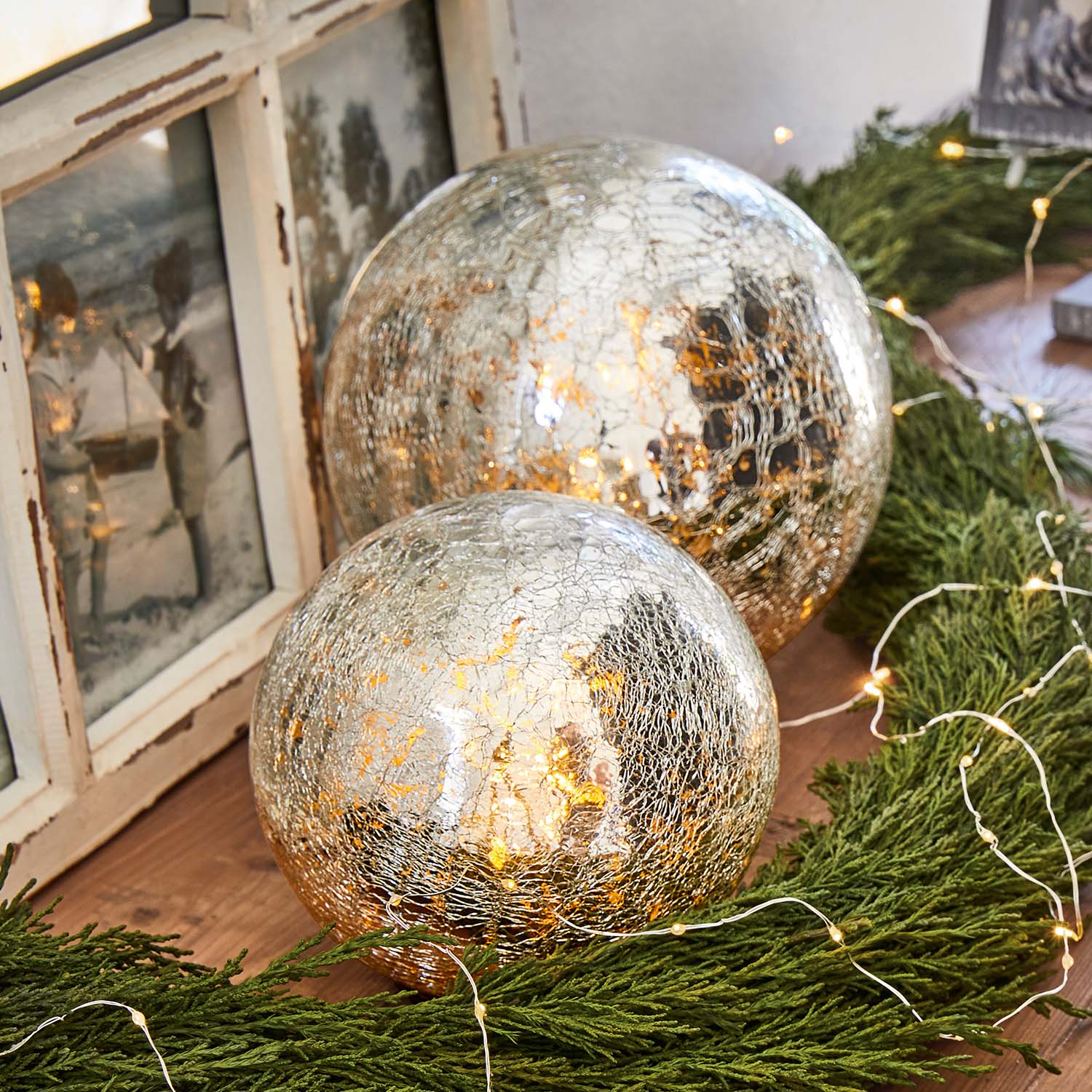 Dos bolas decorativas de vidrio craquelado con luz cálida rodeadas de ramas verdes