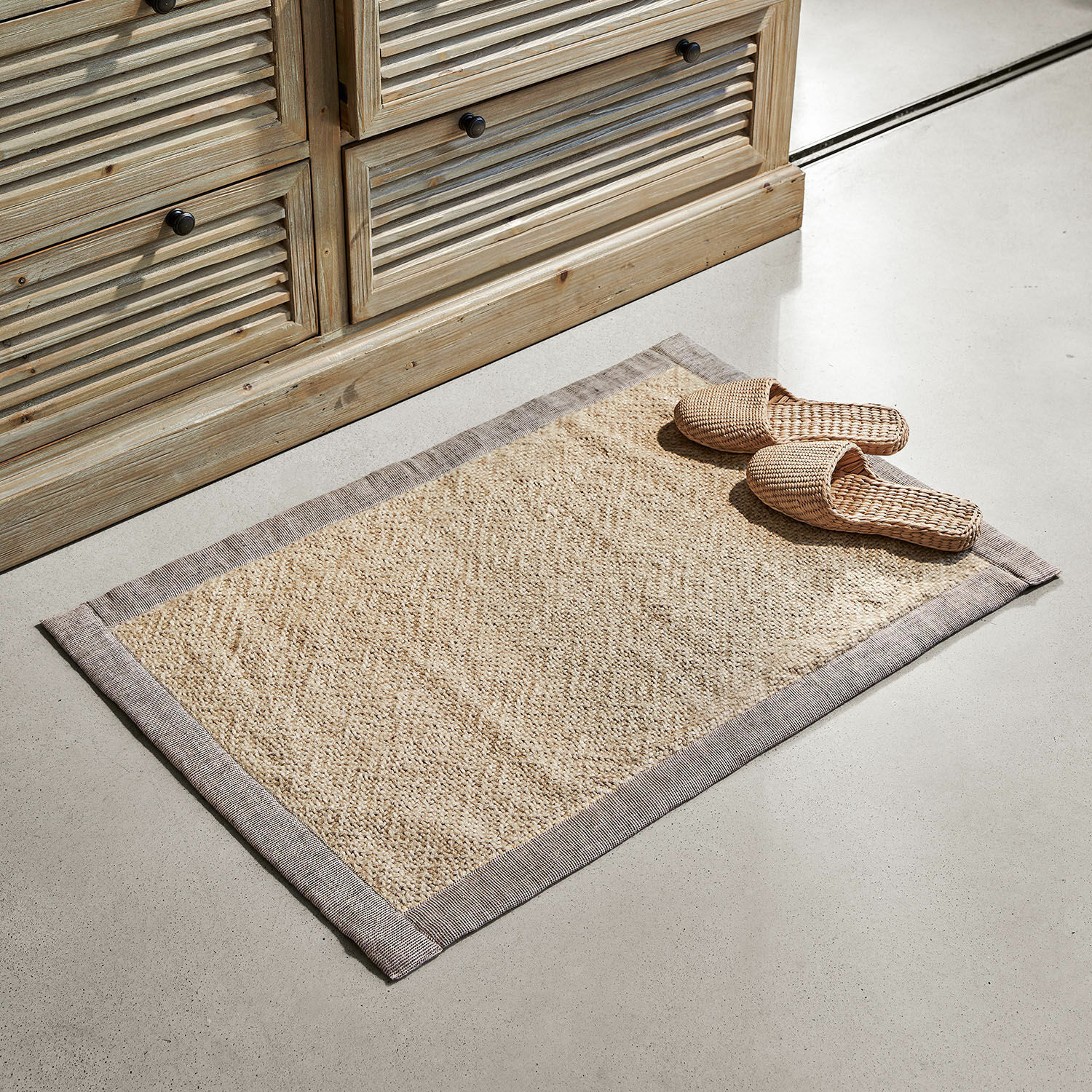 Alfombrilla de baño Wilder Alfombra beige de material natural con borde gris sobre suelo claro junto a mueble de madera