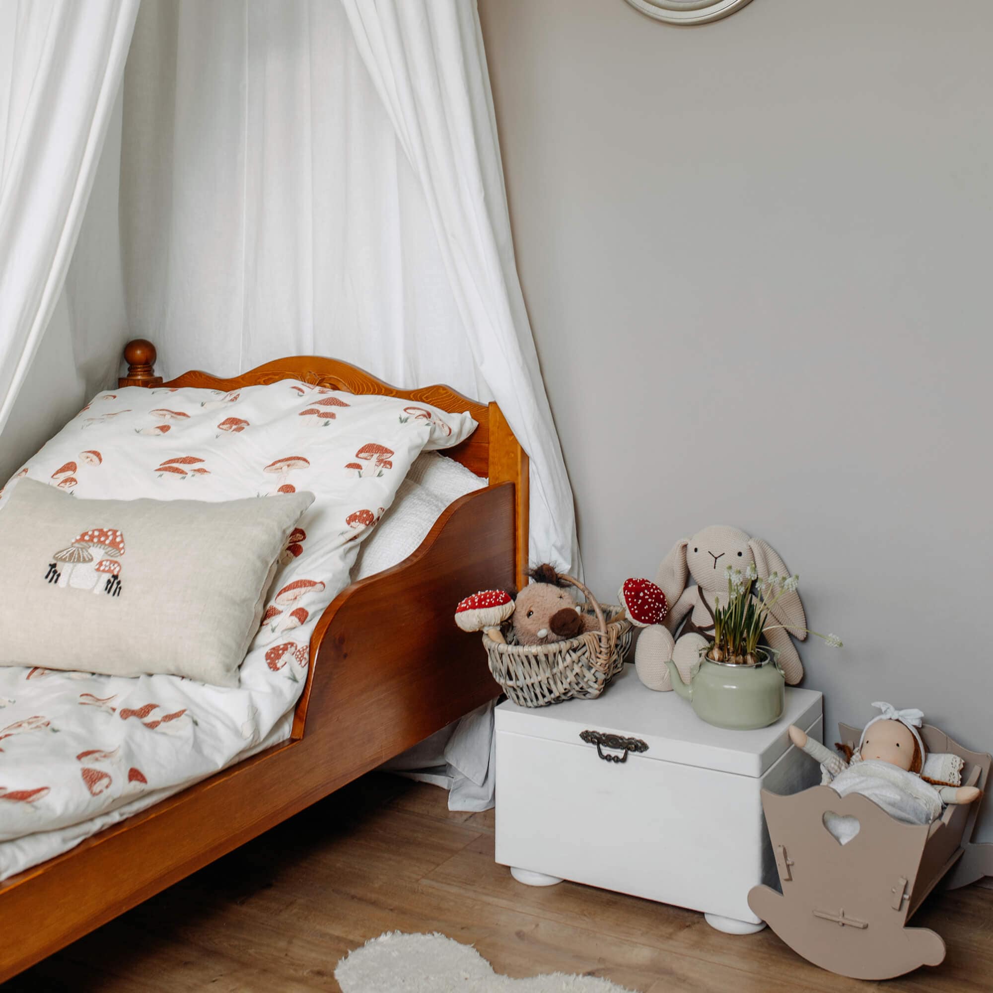 Cama infantil de madera con ropa de cama blanca con estampado de setas y peluches sobre baúl blanco