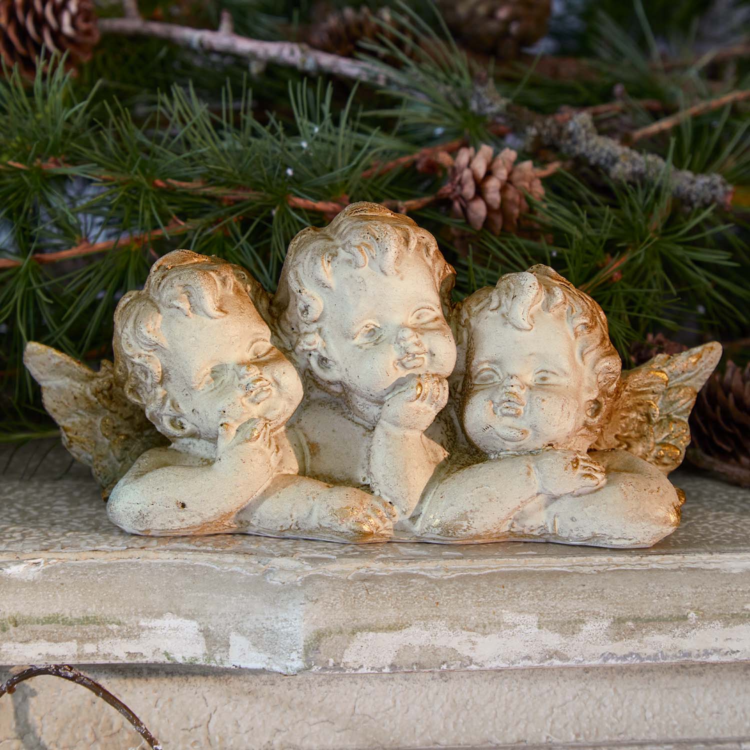Figura de tres ángeles de piedra clara sobre fondo de ramas verdes y piñas, decoración navideña