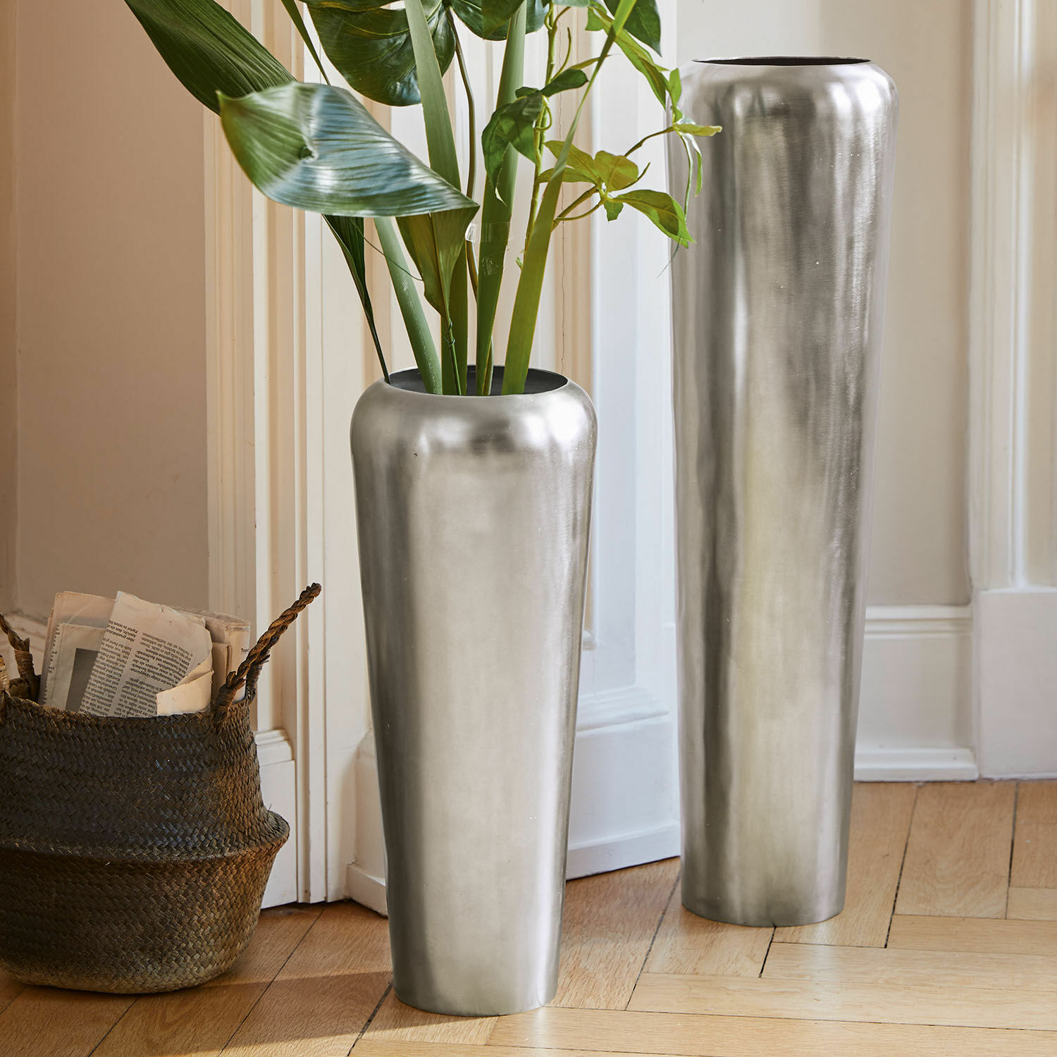 Set de 2 jarrones de suelo Carolan Dos jarrones metálicos altos plateados en el suelo de un interior luminoso con plantas verdes