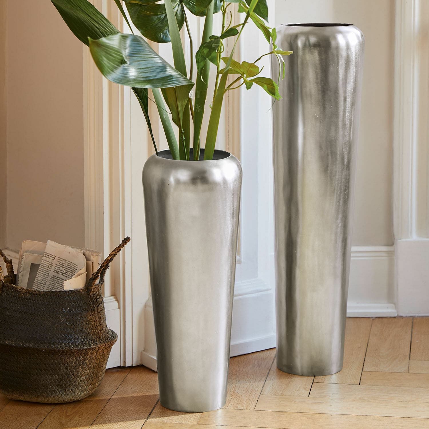 Set de 2 jarrones de suelo Carolan Dos jarrones metálicos altos plateados en el suelo de un interior luminoso con plantas verdes