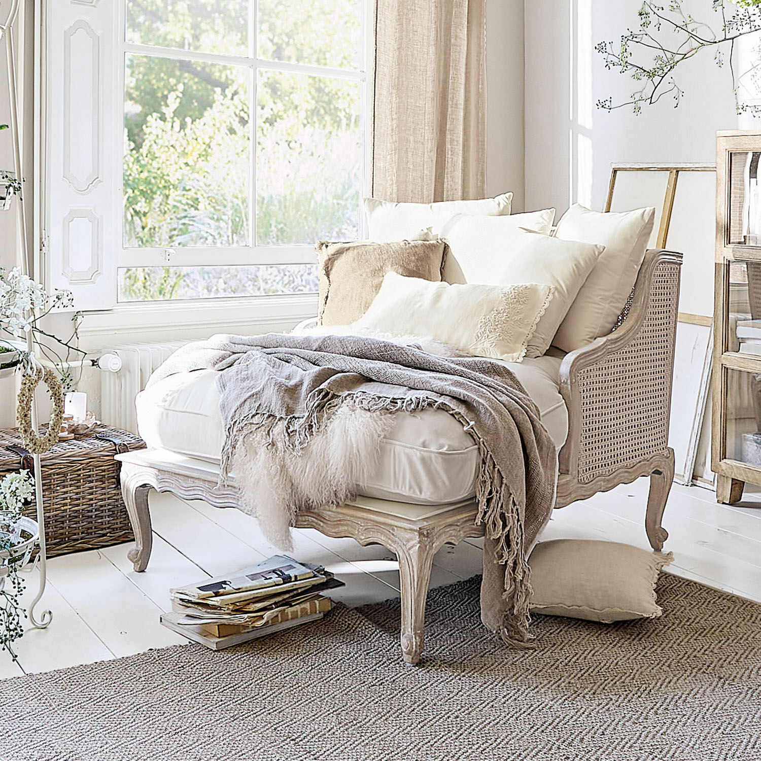 Chaise longue Dronne Sofá beige de madera y ratán con cojines crema y manta suave en interior luminoso