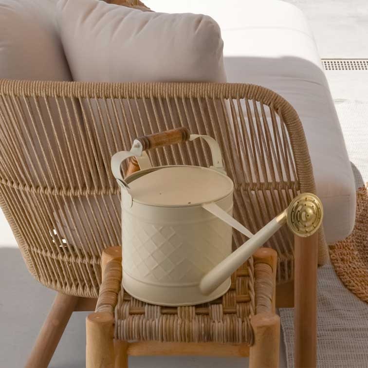 Regadera metálica crema con asa de madera sobre silla de mimbre, decoración de jardín