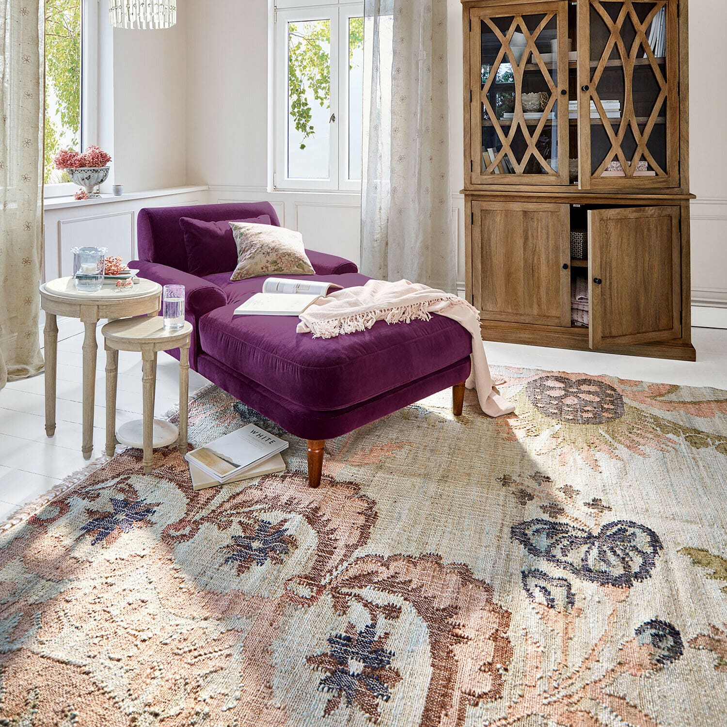 Alfombra Amélorie Chaise longue de terciopelo morado con patas de madera sobre alfombra estampada en salón luminoso con vitrina de madera