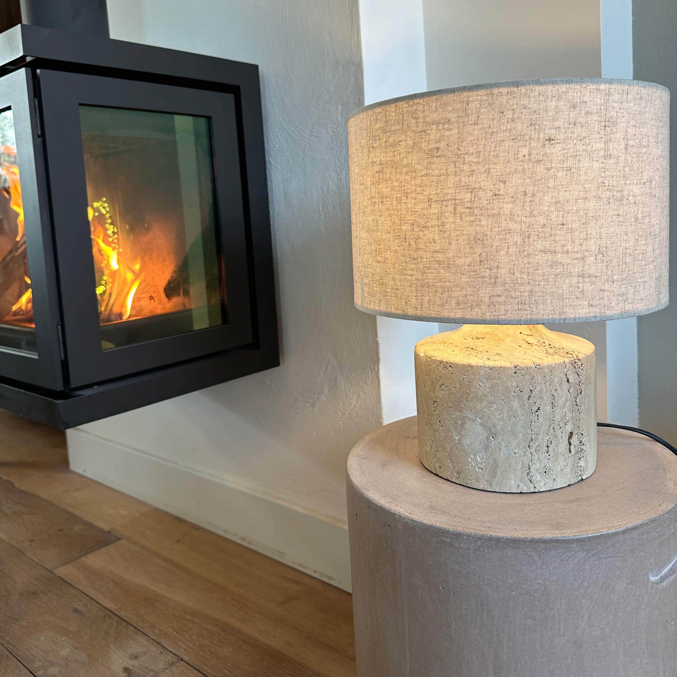 Lámpara de mesa con pantalla beige y base de piedra clara junto a chimenea encendida