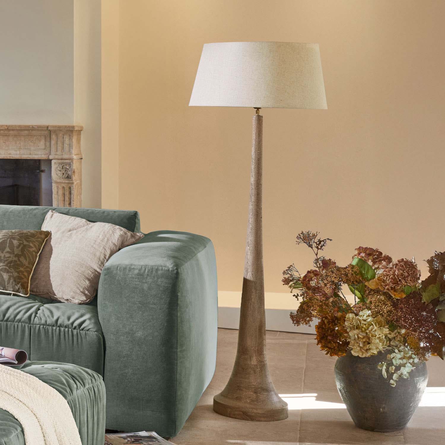 Lámpara de pie con base de madera y pantalla beige en salón moderno con sofá verde y flores secas