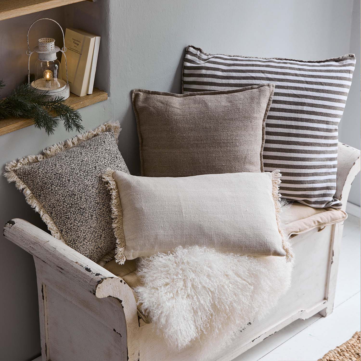 Conjunto de cojines decorativos en tonos beige y marrón sobre banco de madera blanca con manta de piel sintética