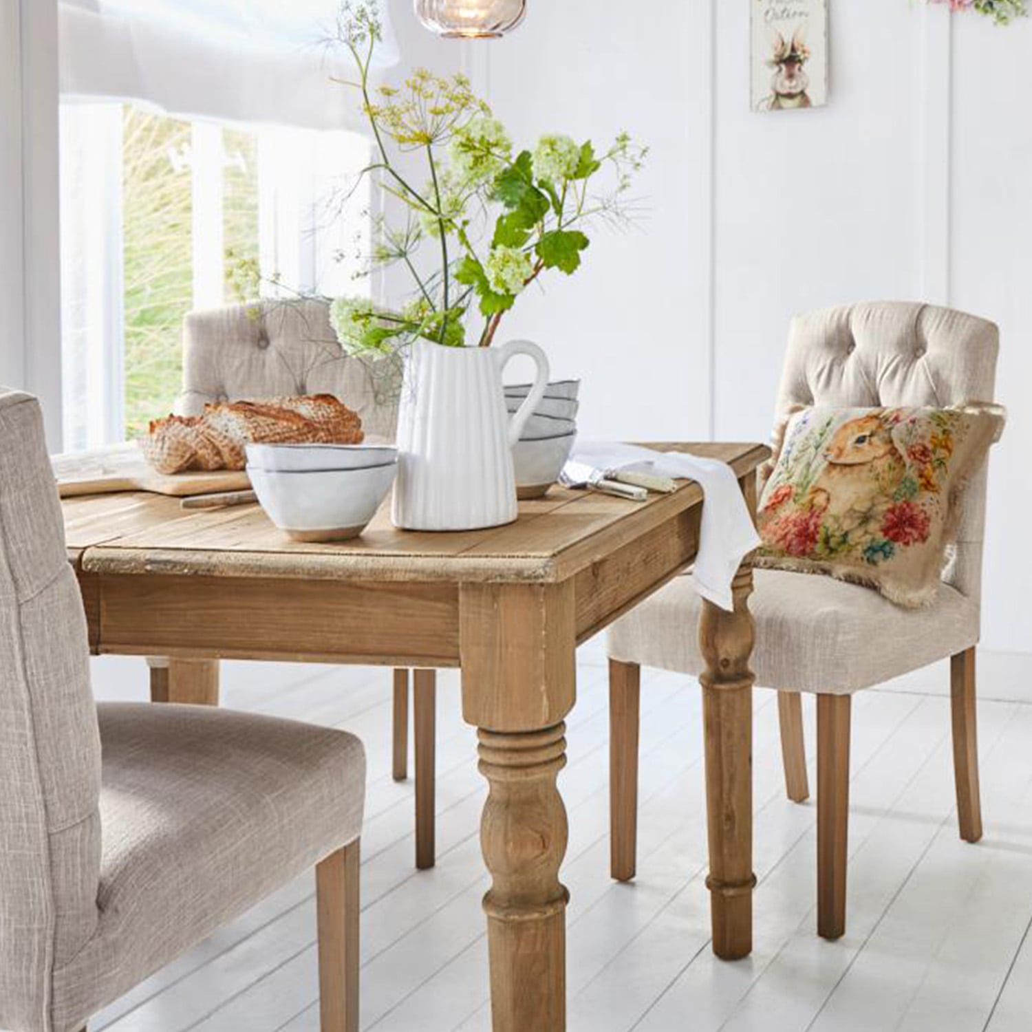 Mesa de madera clara con jarra decorativa y cuencos, sillas tapizadas beige en comedor luminoso